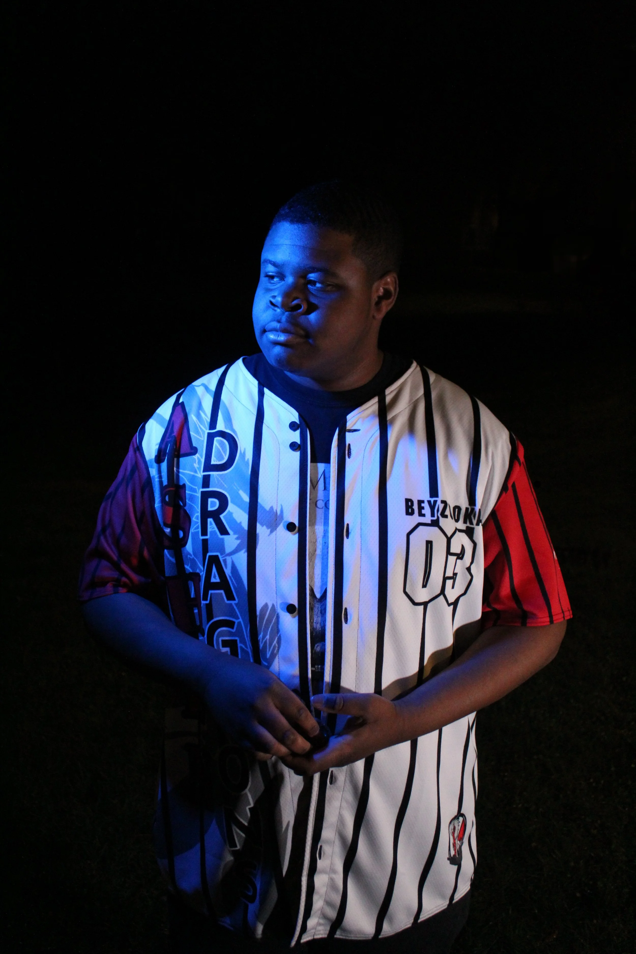 A young man wearing a baseball jersey with vertical black and white stripes, red sleeves, and various printed words and numbers, standing outdoors at night with a dark background and blue lighting on his face and clothes.