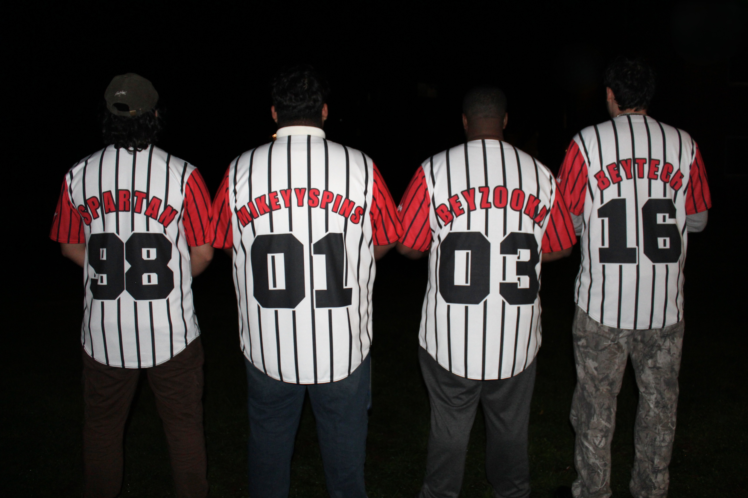 Four people standing with their backs to the camera, all wearing matching striped baseball jerseys with names and numbers on the back, in a dark setting.
