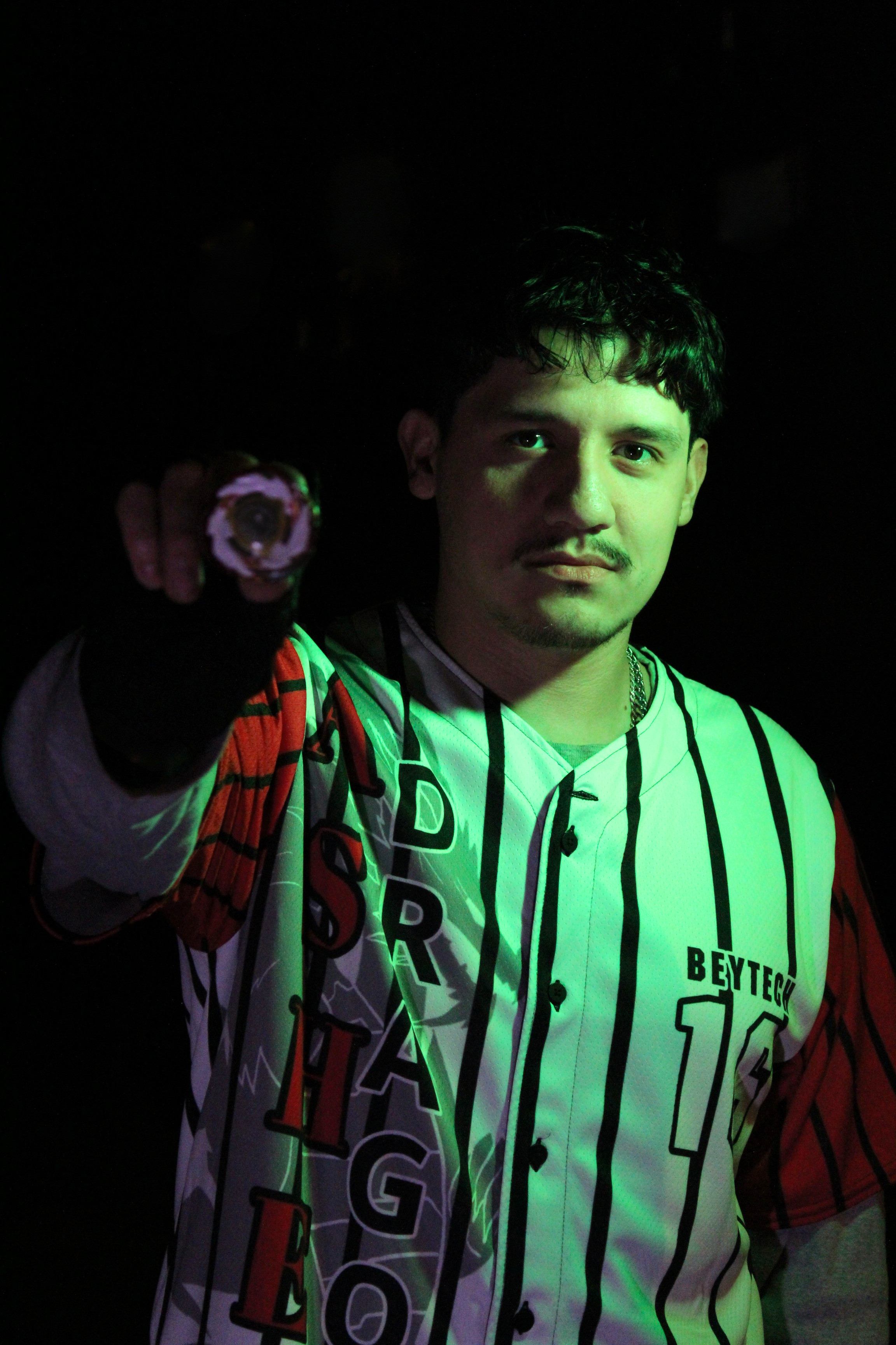 Young man with dark hair and light beard wearing a baseball jersey and black glove, pointing forward, against a dark background with green lighting.