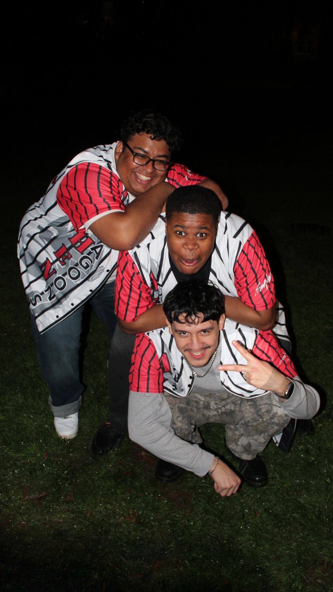 Three young men in matching baseball jerseys pose together outdoors at night, smiling and making playful gestures.