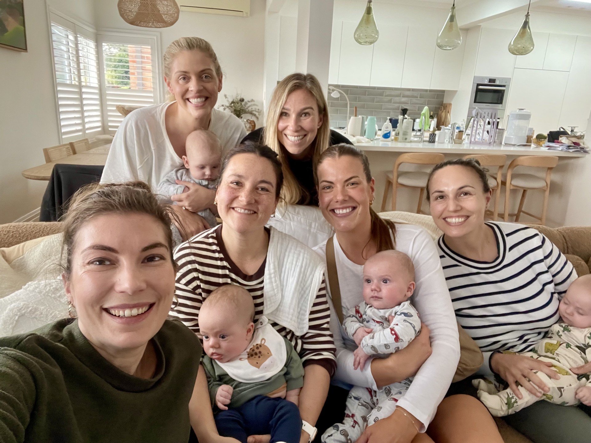 A group of women and children gathered in a living room, smiling and taking a selfie. The background shows a kitchen with white cabinets and a dining area with wooden chairs.