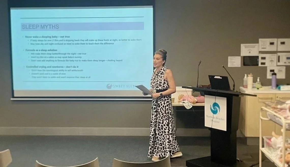 A woman in a leopard print dress giving a presentation in a classroom or seminar setting. A screen behind her displays a slide titled 'Sleep Myths'. There is a lectern with Northern Beaches Hospital logo to her right and medical supplies to the side
