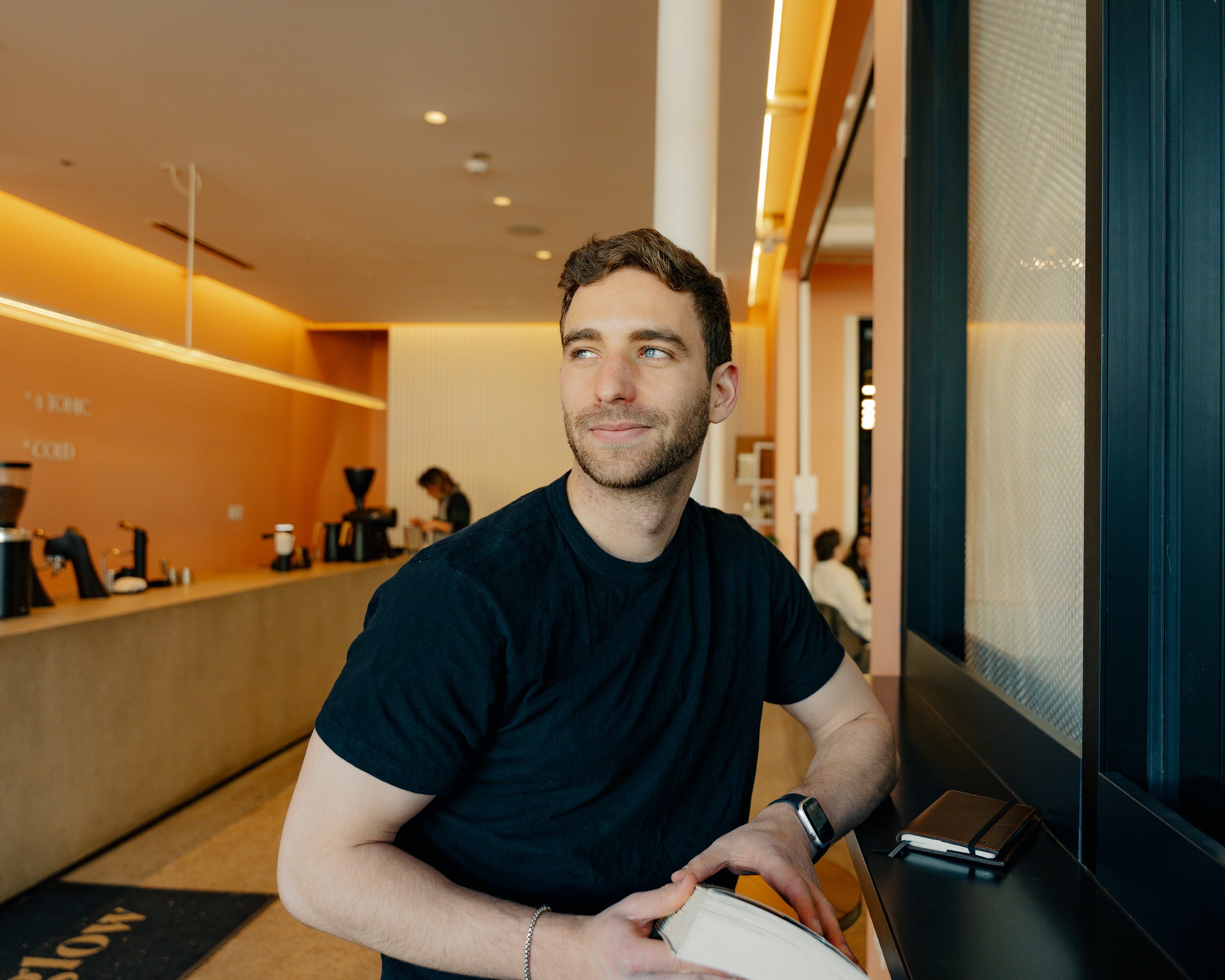 A young man with short dark hair and a beard, wearing a black t-shirt and a smartwatch, sitting at a counter in a modern, warmly lit cafe or restaurant with orange and beige walls, and looking sideways against a dark wall panel.