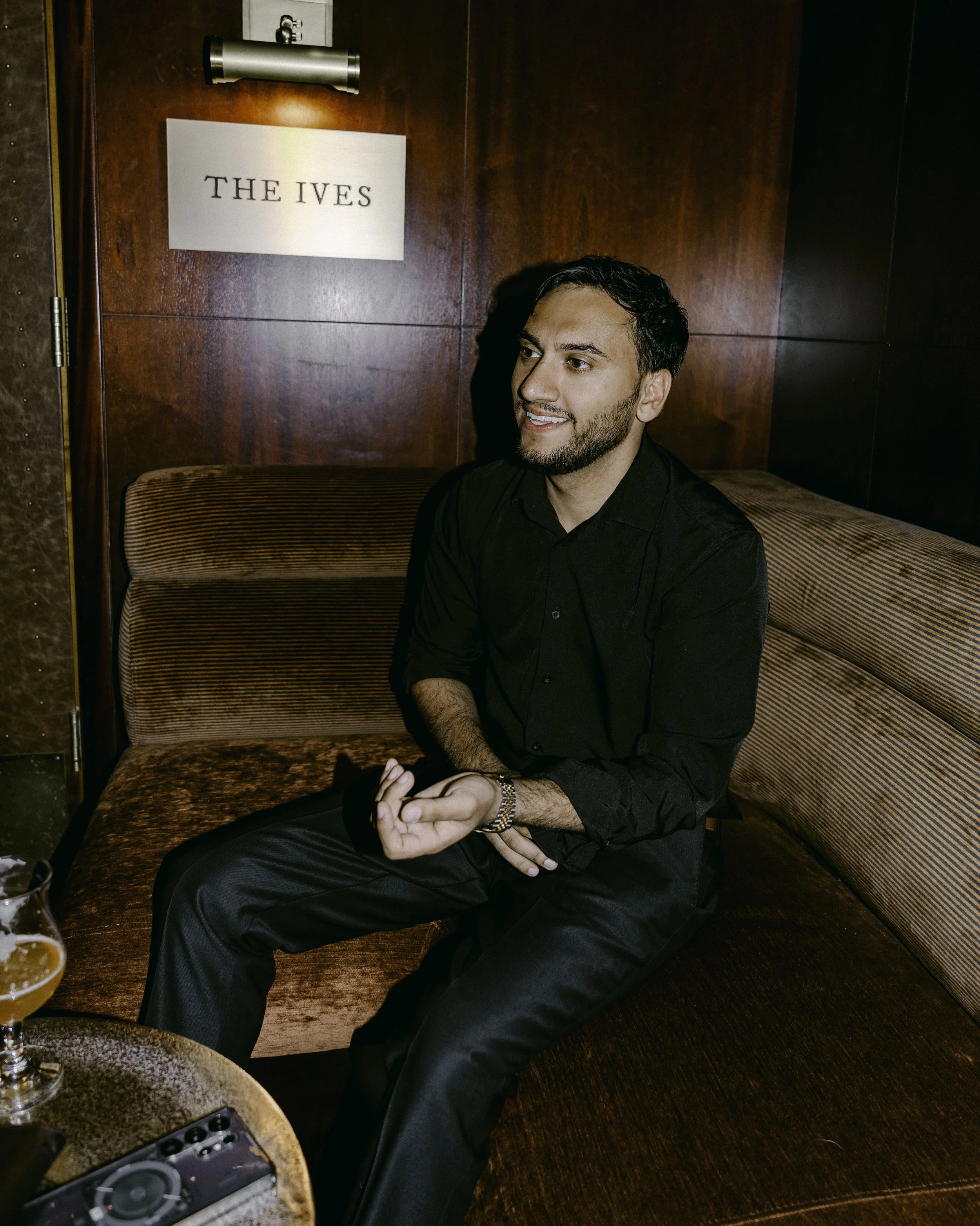 A man with dark hair and a beard, wearing a black shirt, sitting on a curved, striped, brown velvet bench in a wine bar. There is a sign reading 'THE IVES' on the wooden wall behind him. A drink and a DJ controller are on the table in front of him.