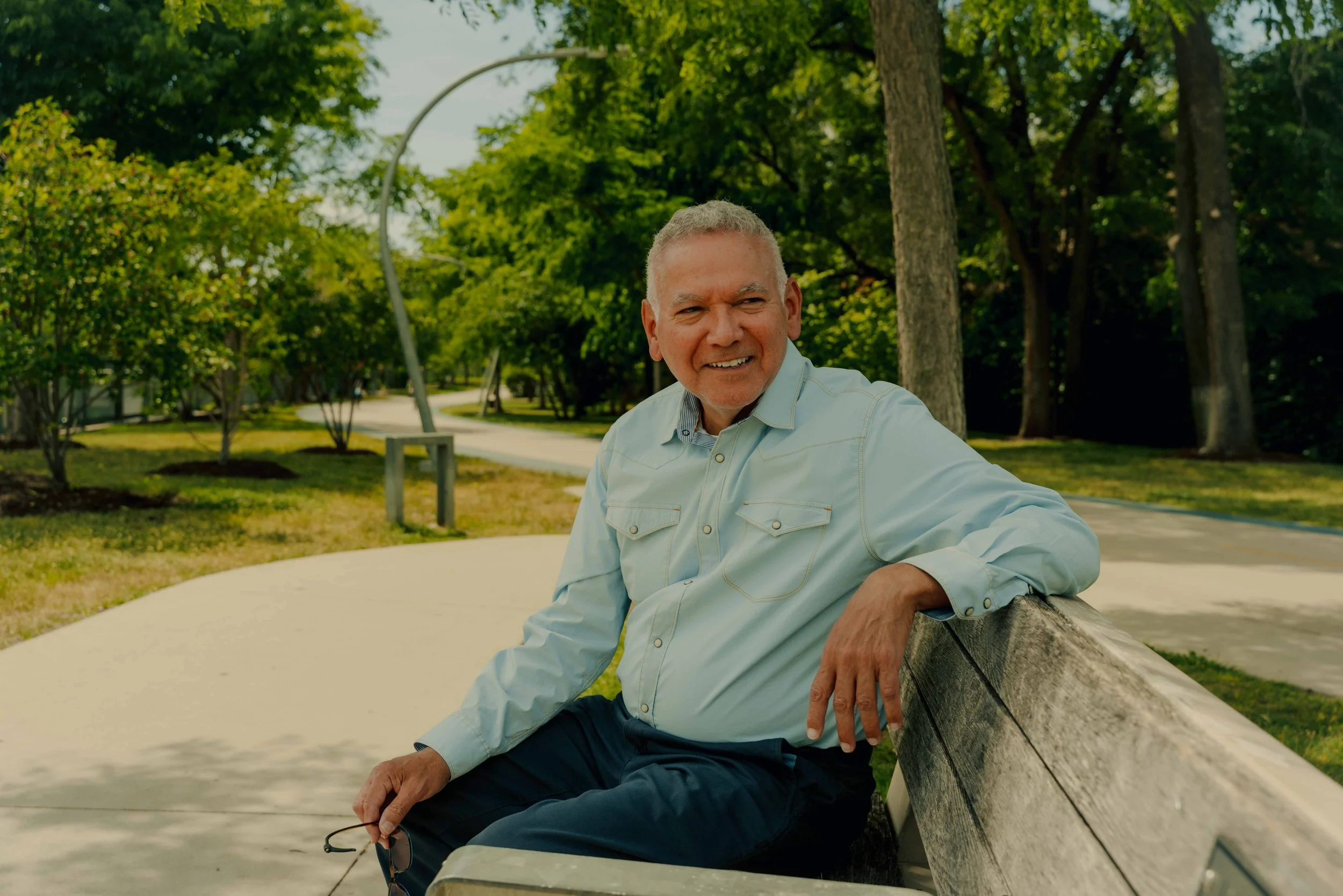 An older man with gray hair sitting on a park bench, smiling, holding glasses, surrounded by green trees on a sunny day.