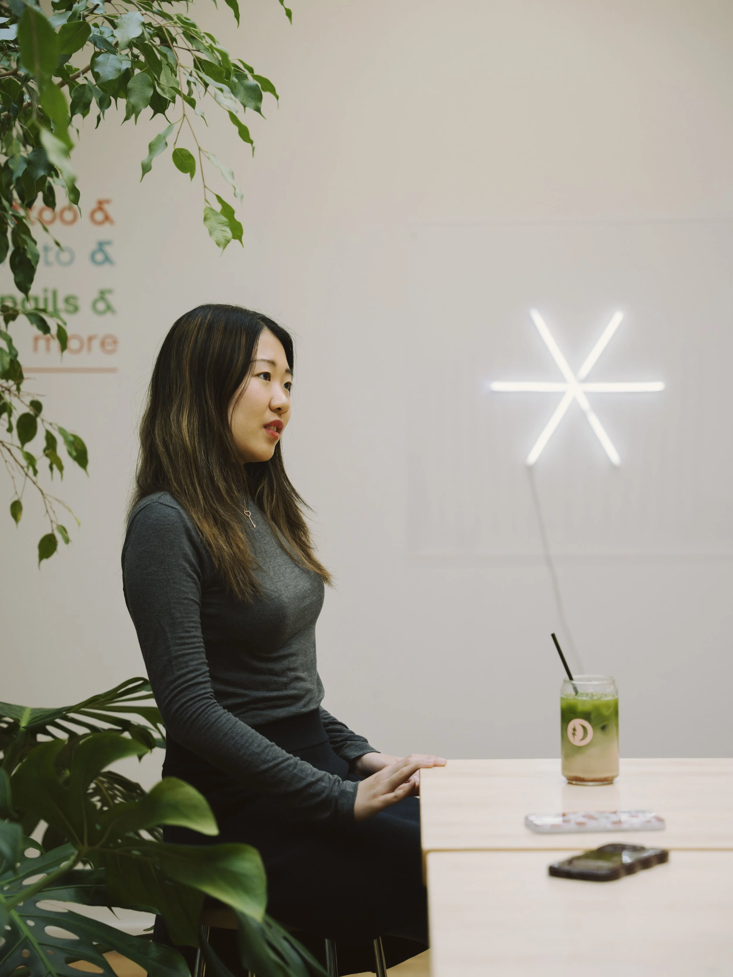A woman with long dark hair, wearing a dark gray long-sleeve shirt, sitting at a light-colored table in a modern, minimalistic space with white walls and green plants. There is a colorful drink with a straw and a ghost face sticker on the glass on th