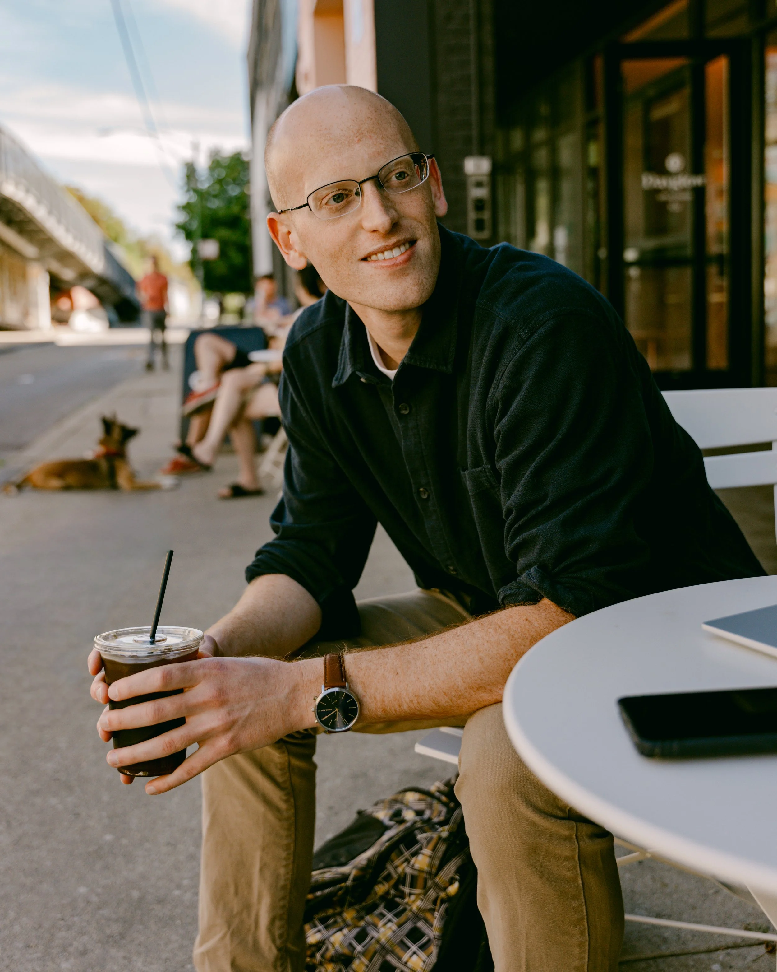 A person sitting at an outdoor table holding a cold drink with a straw, wearing a watch and tan pants, with other people in the background on a city street.