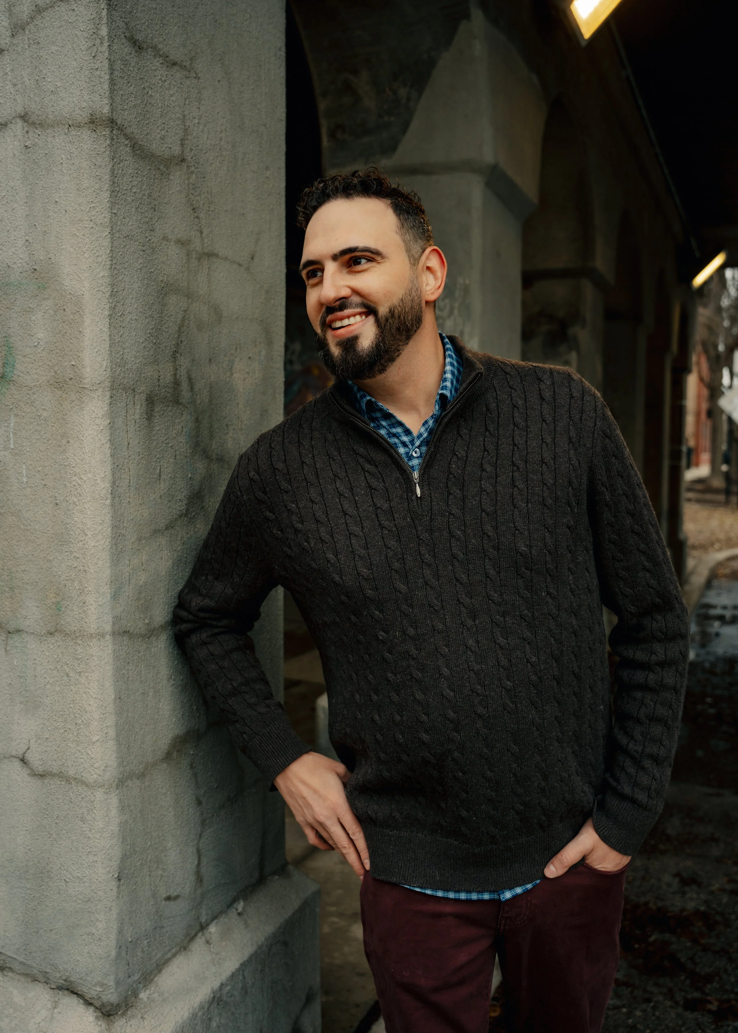 A man with dark curly hair, beard, and light skin standing outdoors near a concrete wall, smiling and casually resting his left hand on his hip, wearing a dark knit sweater over a blue checkered shirt with maroon pants.
