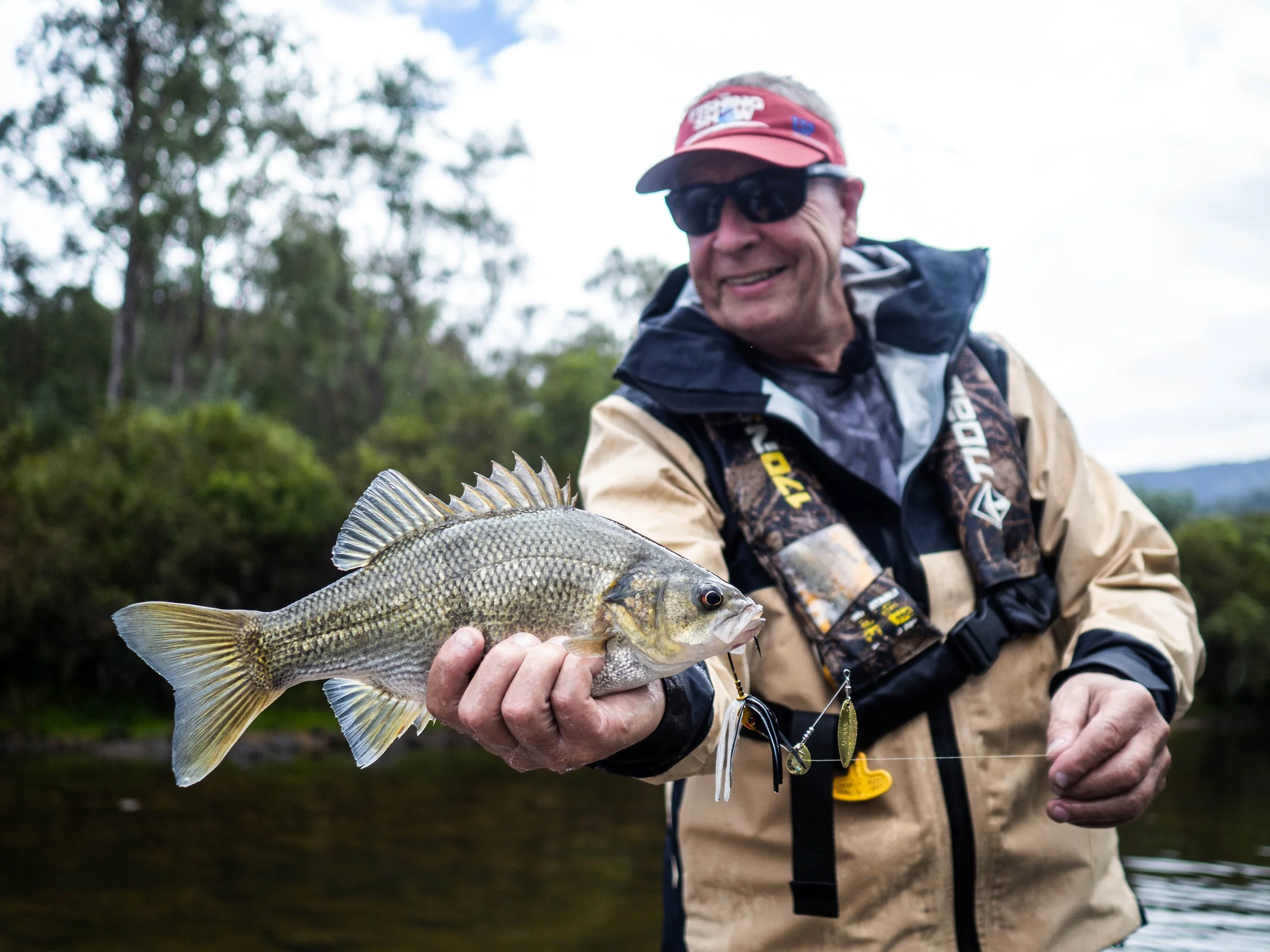 An older man wearing sunglasses, a red cap, and a beige and black jacket holding a fish on a fishing line during daytime outdoors.