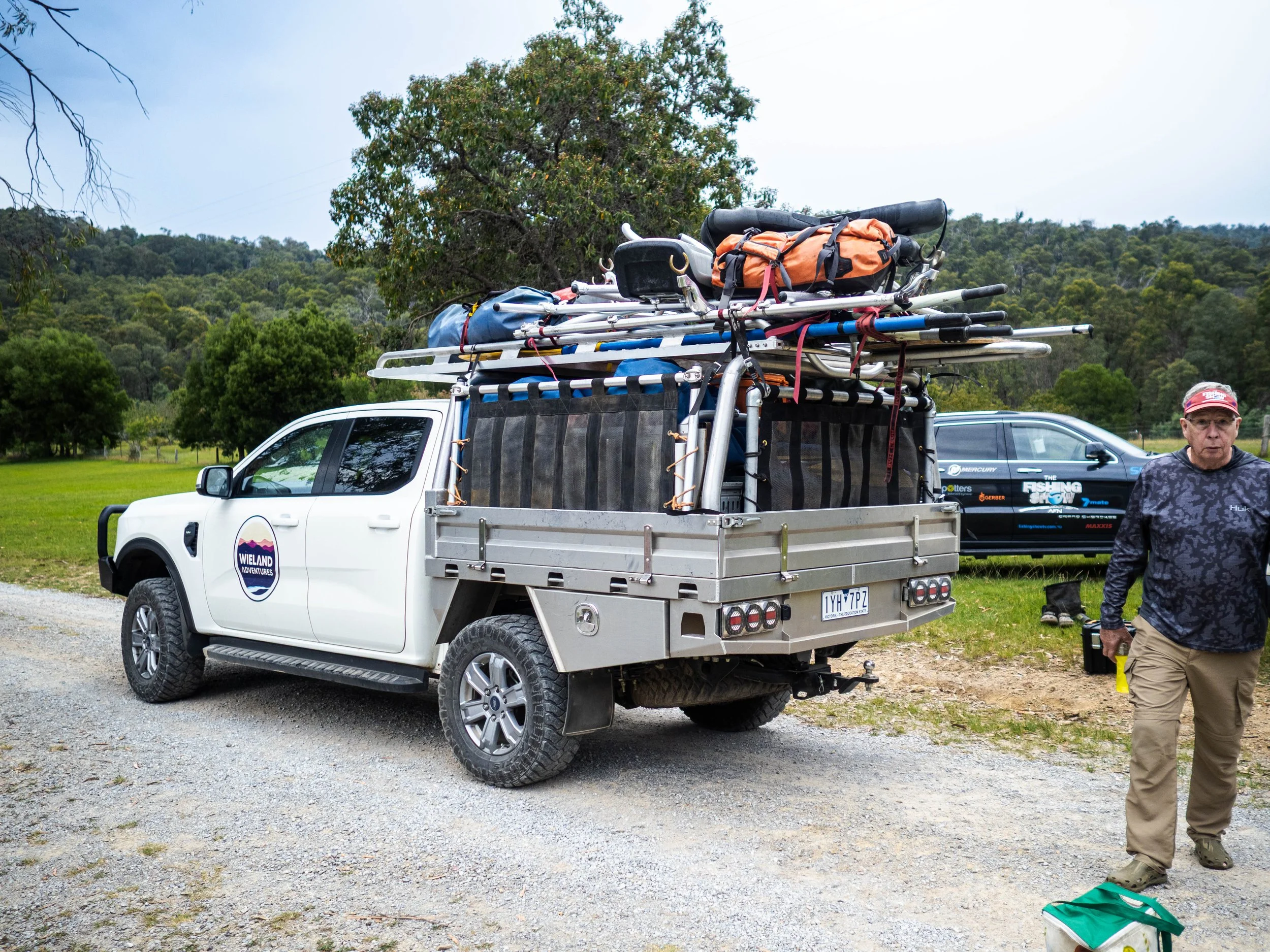 White pickup truck loaded with camping and outdoor gear parked on a gravel area in a park with trees and grassy fields, with a man standing nearby.