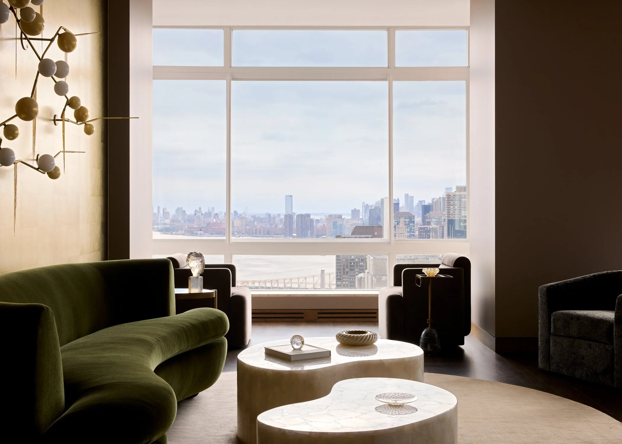 Living room with large window overlooking a city skyline, green velvet sofa, black armchair, small marble coffee tables, and decorative items.