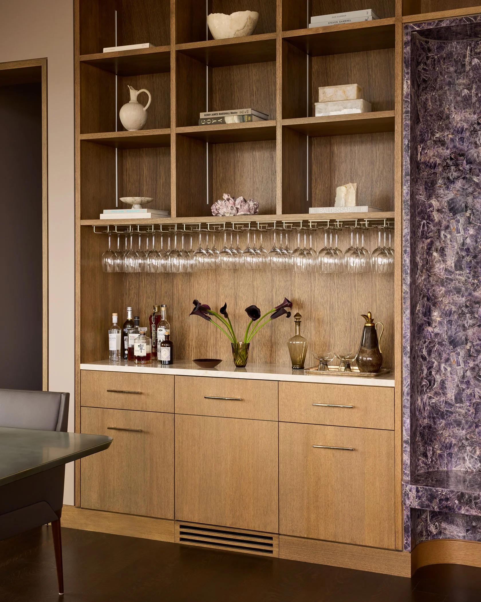 Wooden bar cabinet with glassware, liquor bottles, and decorative items, next to a purple marble wall.