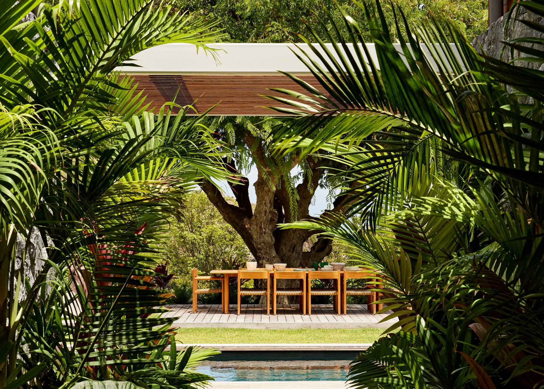 Outdoor patio with a large man-made tree, surrounded by lush tropical plants and palm leaves, with a wooden dining table and chairs on a wooden deck near a swimming pool.