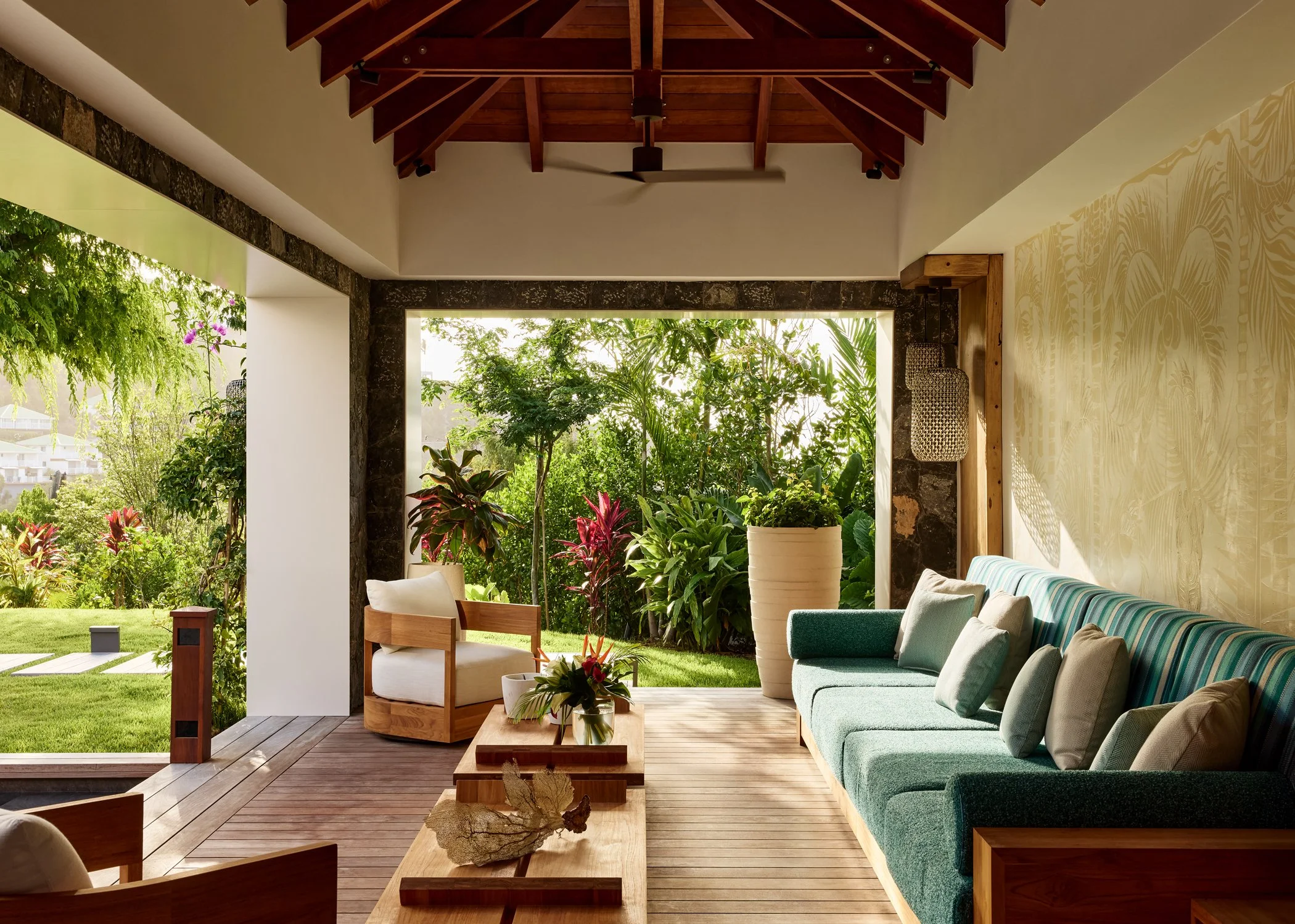 Living room with open view of lush green garden, wooden furniture including a sofa with cushions, and potted plants, with sunlight streaming in.