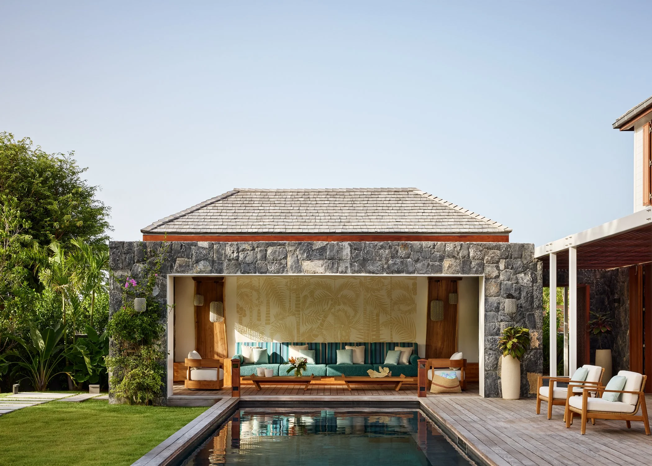 Outdoor patio with a pool, cushioned seating area, and lush greenery under a clear blue sky.