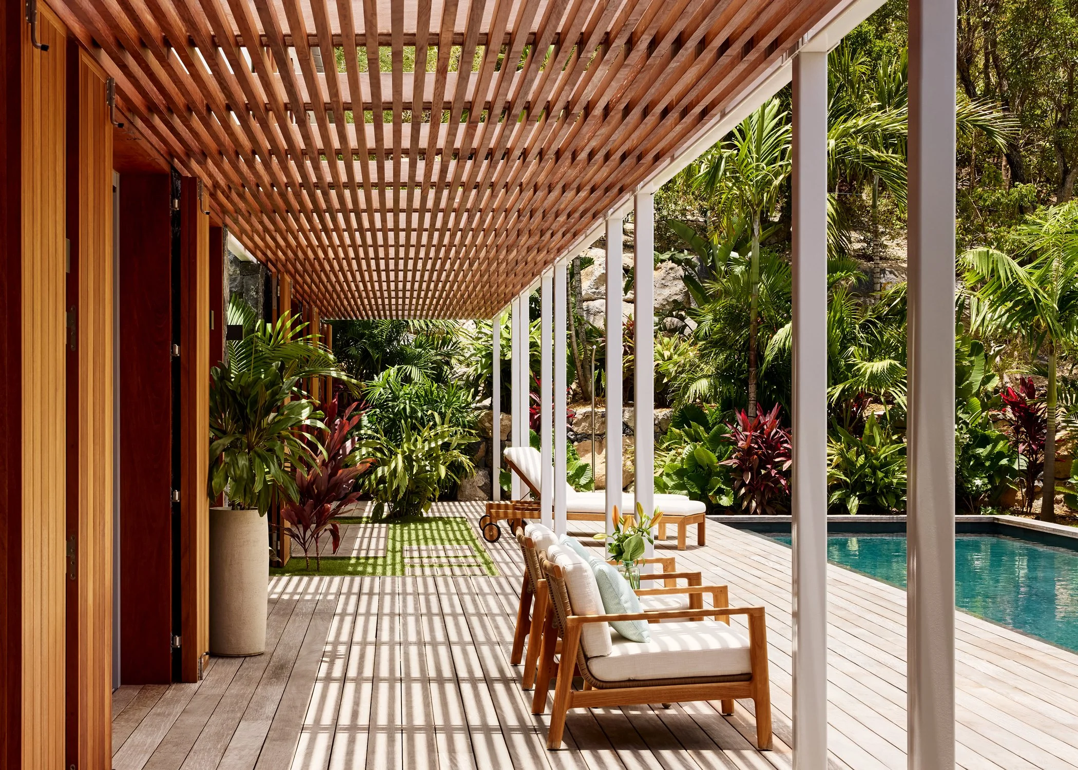A modern outdoor patio area next to a swimming pool, featuring wooden chairs with cushions, lush tropical plants, and a wooden pergola providing partial shade.