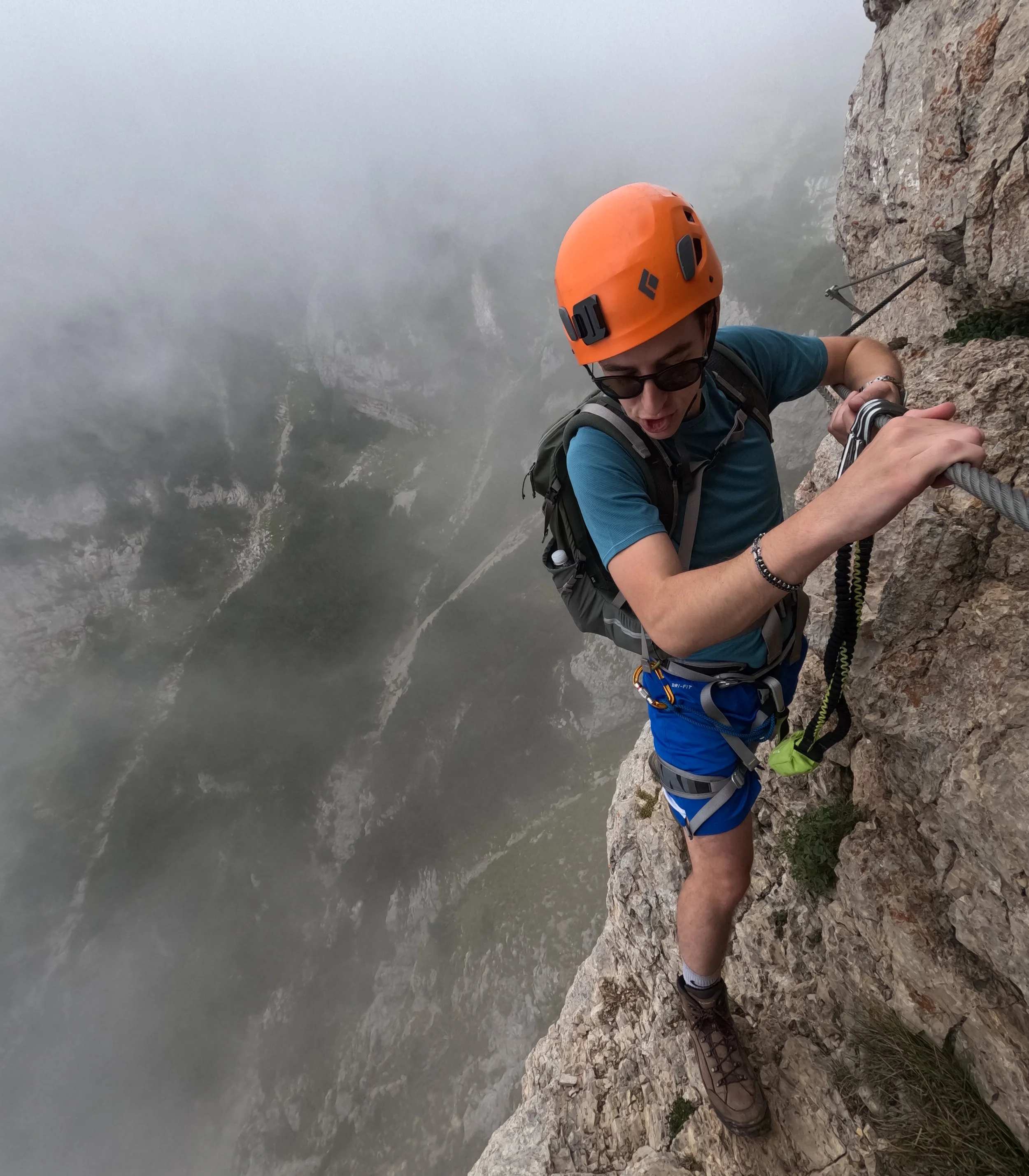 Davide Moro con casco arancione Black Diamond indossa un equipaggiamento da arrampicata mentre scala una parete rocciosa sulla cima di una montagna, con una vista di una valle e nebbia sottostante.