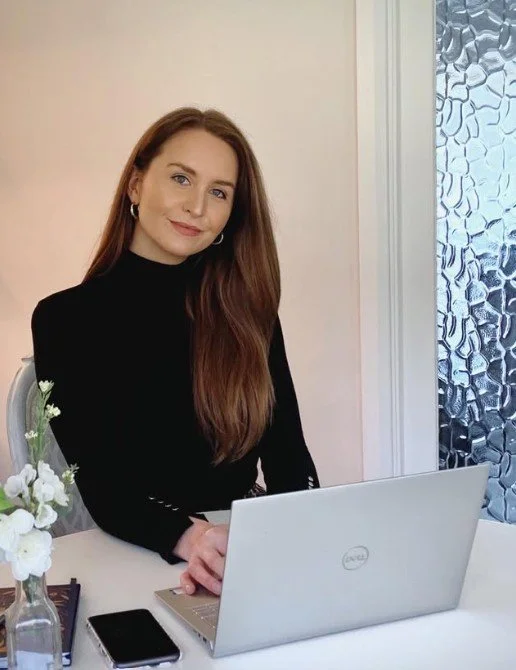 A woman with long, straight, reddish-brown hair sitting at a white desk with a laptop, a smartphone, a book, and a small vase of white flowers, in front of a textured glass window.