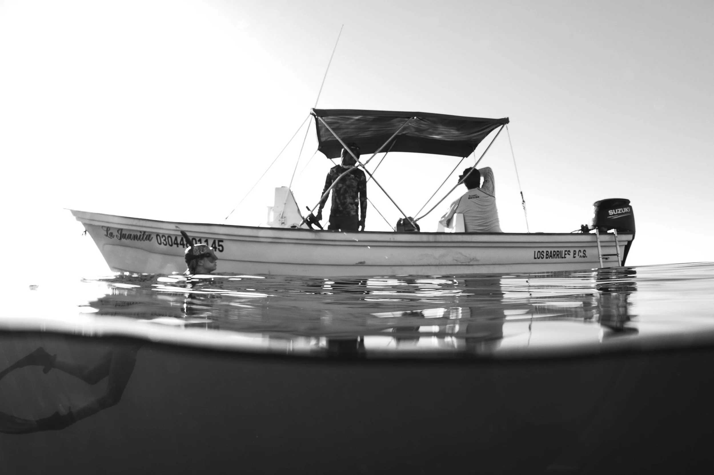 Black and white over-under split photo of a small dive boat on the surface with a swimmer below the waterline.