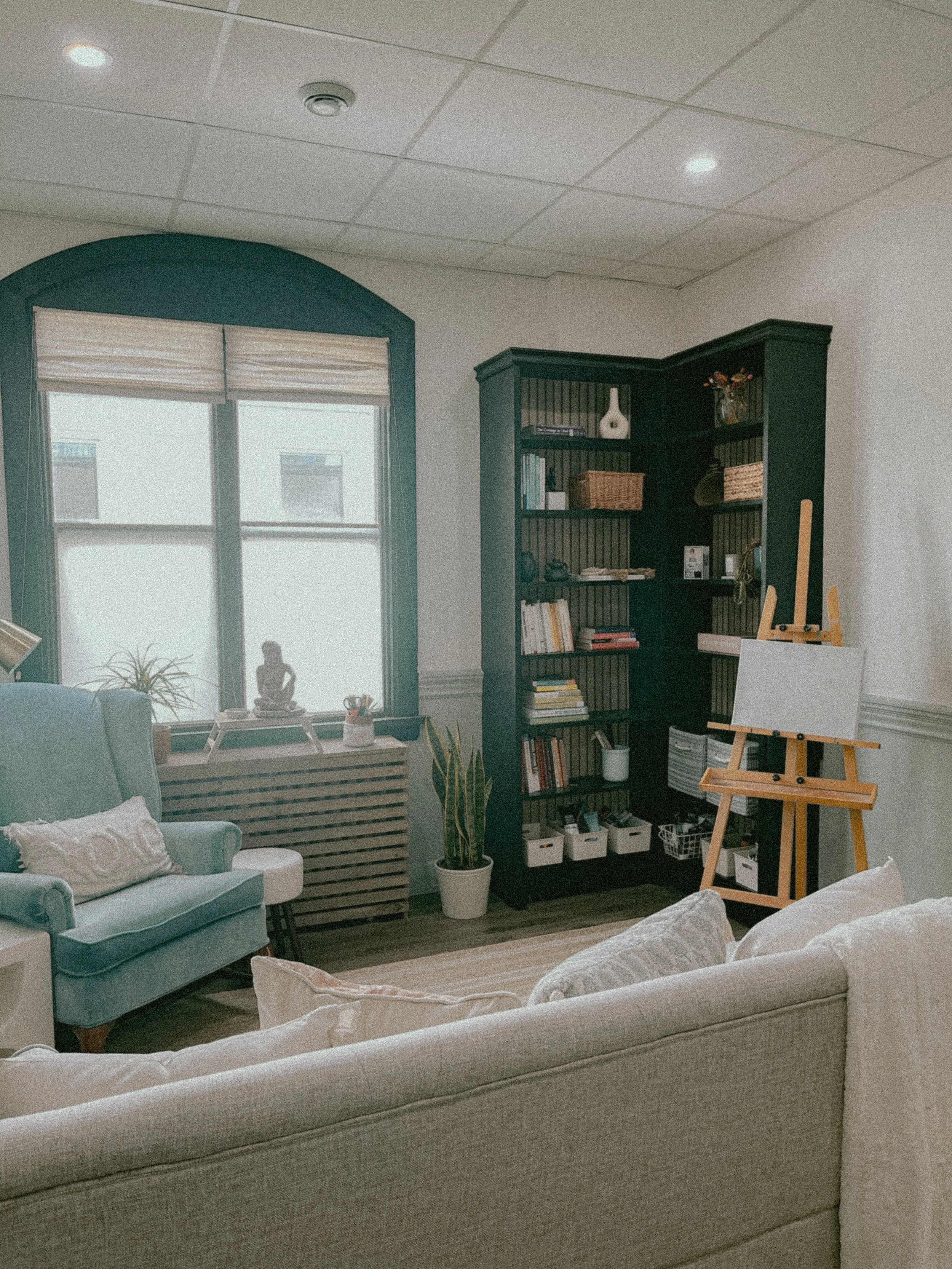 Living room with beige sofa, armchair, bookshelf, window with blinds, potted plants, and an easel.