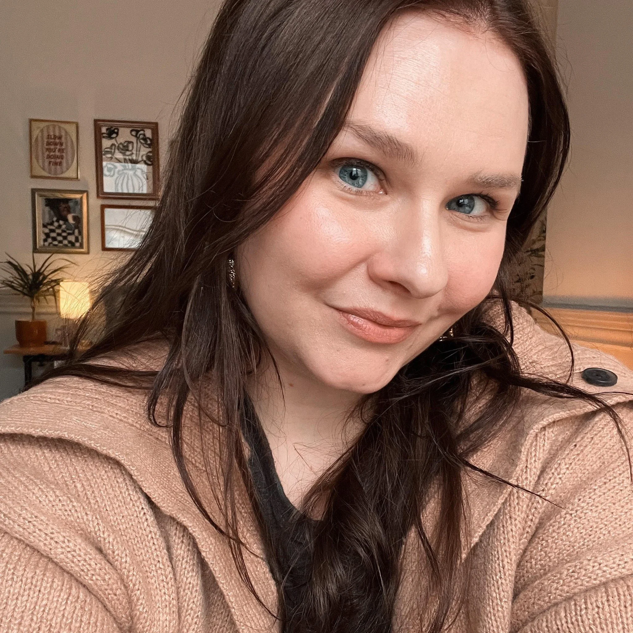 Close-up of a woman with blue eyes, dark brown hair, and wearing a peach-colored sweater, standing in a room with framed pictures on the wall and a lamp in the background.