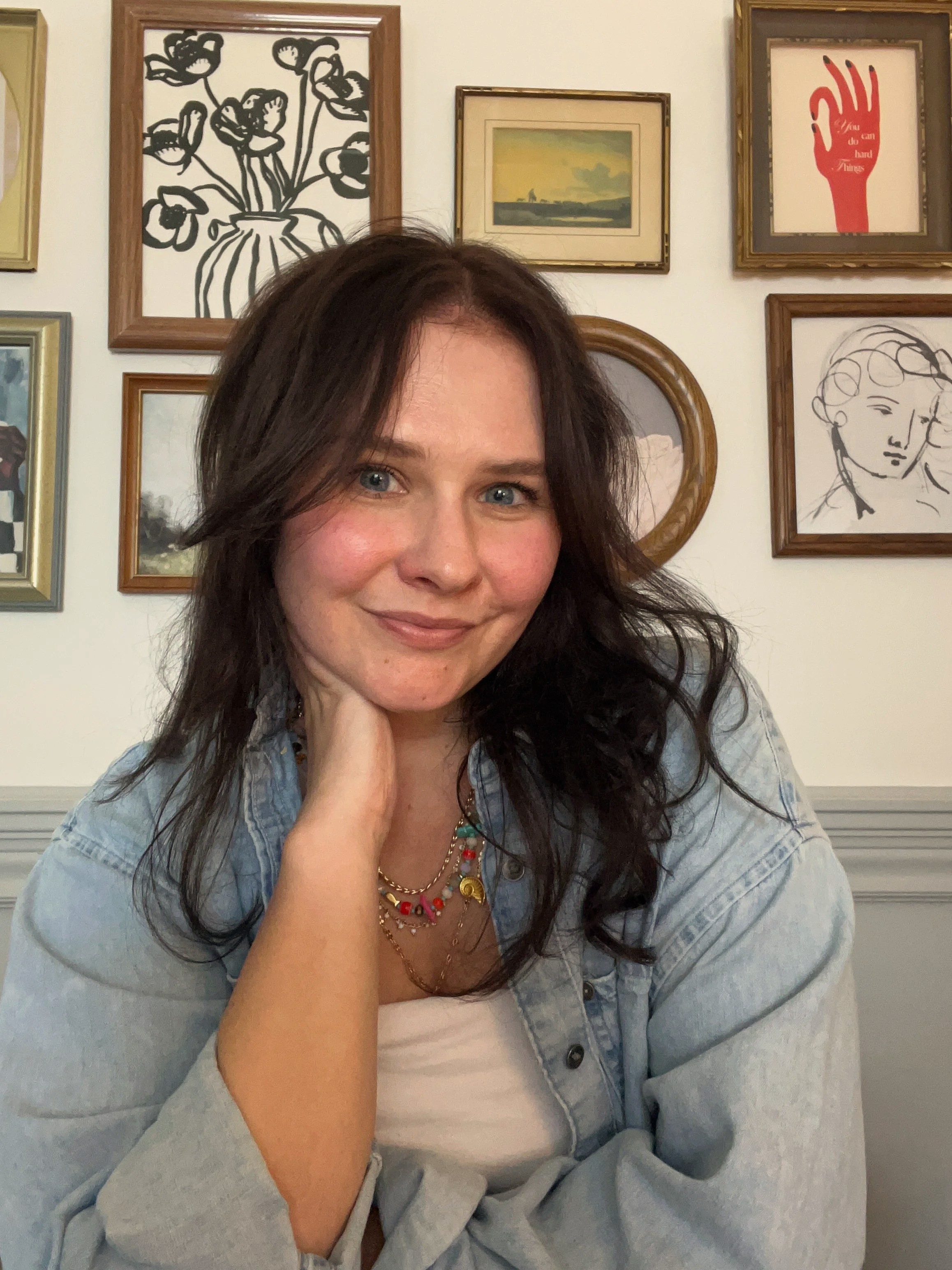 A woman with long dark hair and blue eyes smiling at the camera in a room with multiple framed artworks on the wall behind her, including a black and white flower drawing, a landscape painting, and a red hand illustration.