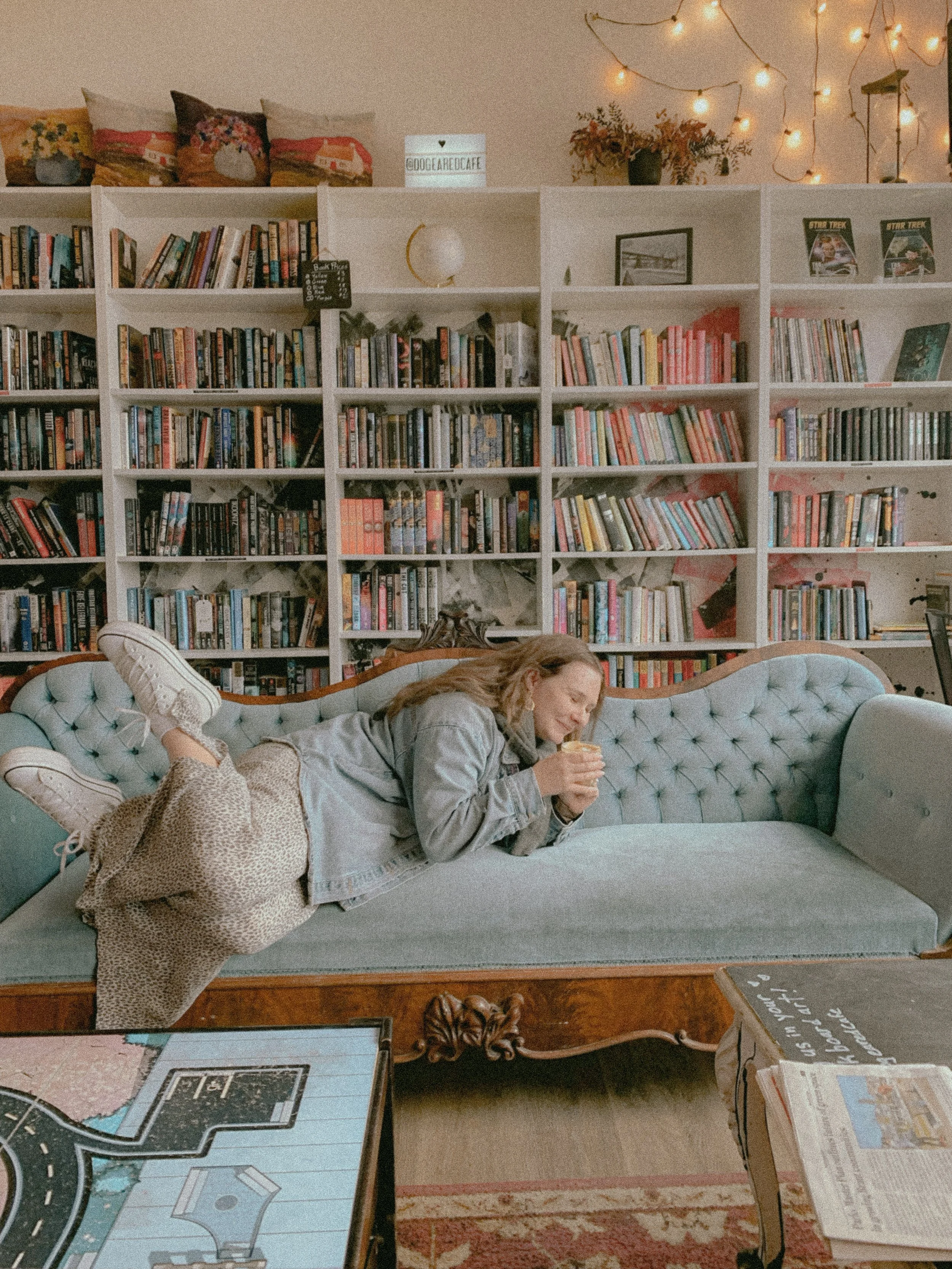 A woman with curly hair lying on a vintage-style couch in a cozy, well-decorated living room, holding a glass of drink, with a large white bookshelf filled with books and framed pictures behind her, and warm string lights on the wall.