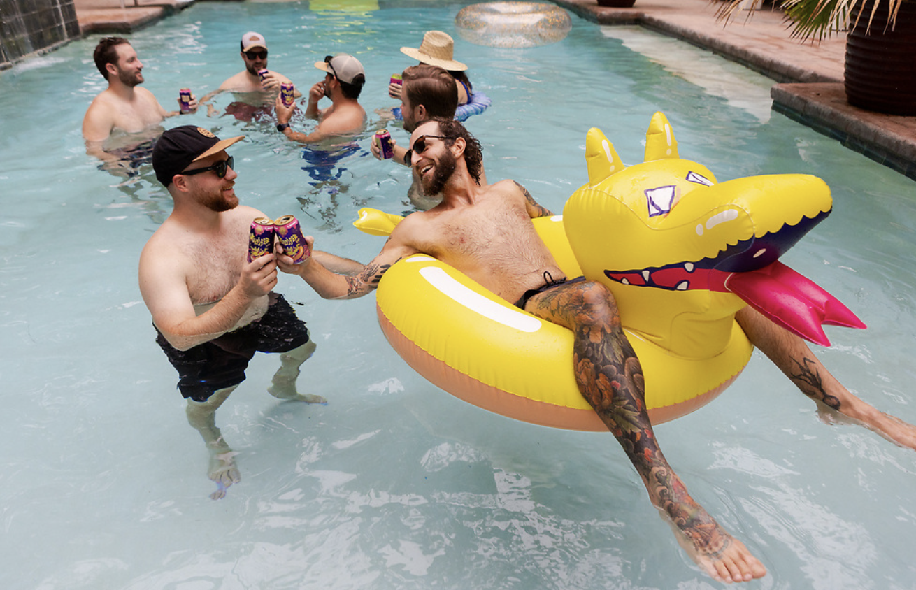 Two men smile with their beers in a pool. One is in a yellow float