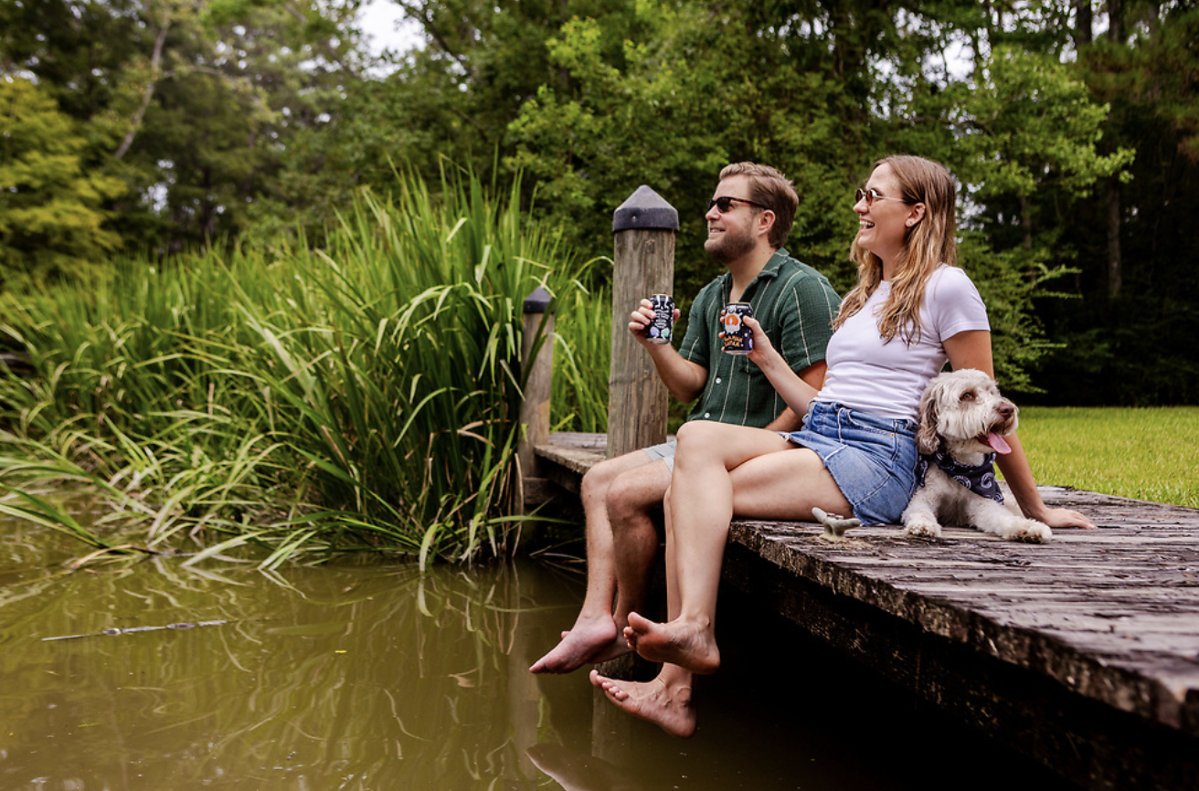 A couple sits with their dog on a dock drinking a beer