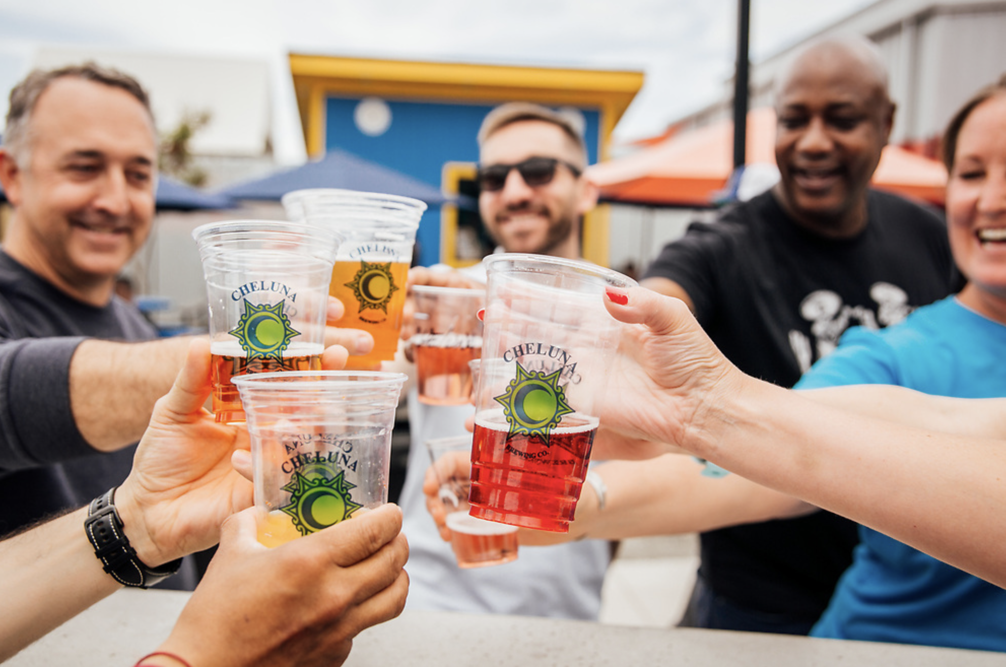A group of 7 adults toast their beers in cheluna brewing co. cups
