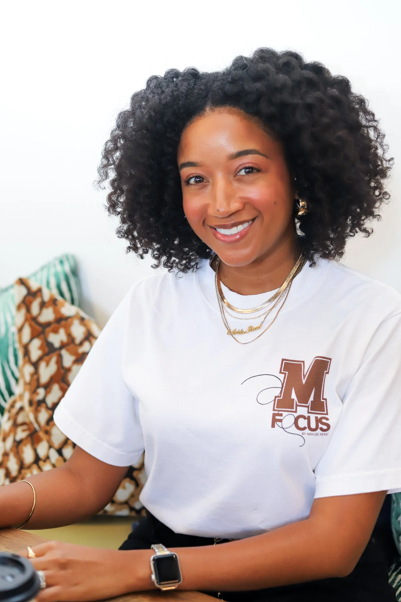 A smiling woman with curly hair wearing a white t-shirt with a logo on it, sitting at a table with a watch and jewelry, and a patterned bag in the background.