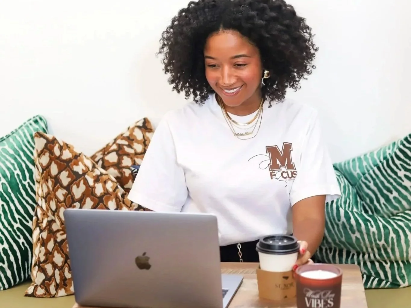 A woman sitting on a sofa with patterned pillows, working on a silver MacBook laptop, holding a coffee cup, wearing a white t-shirt with an 'M Focus' logo, smiling while looking at her laptop.