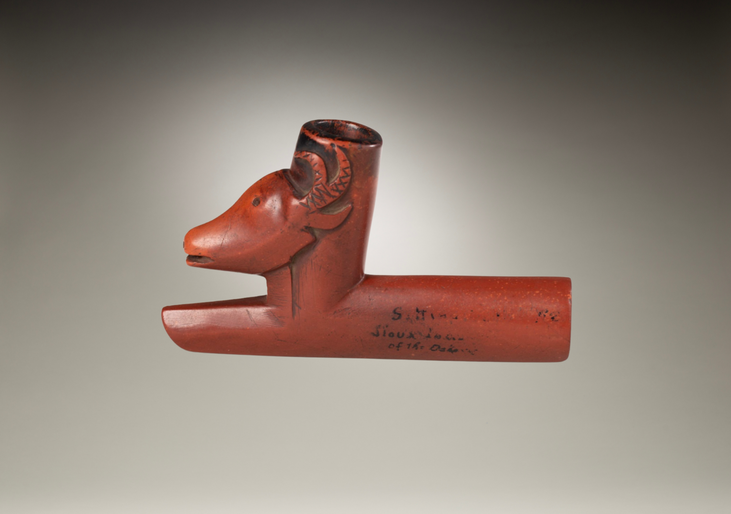 A red-orange ceramic art piece shaped like a stylized animal, possibly a horse or dog, with a rounded head, a short snout, and decorative carvings on the face and neck.