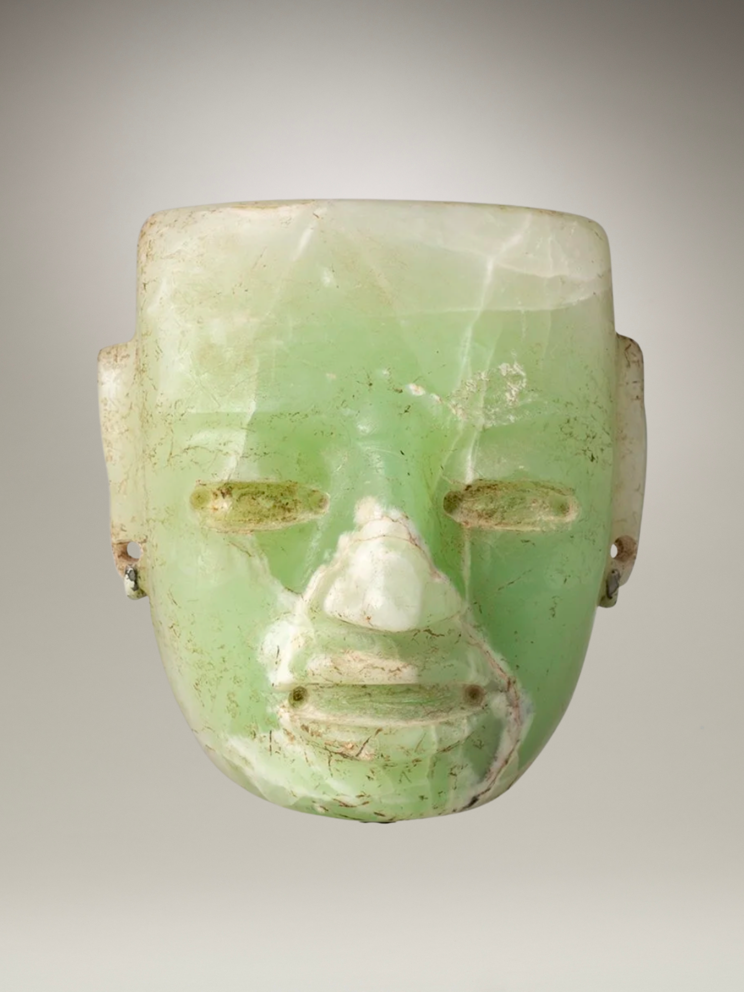A carved jade mask with a pale green color, intricate facial features, and holes for eyes and nostrils, displayed against a plain background.