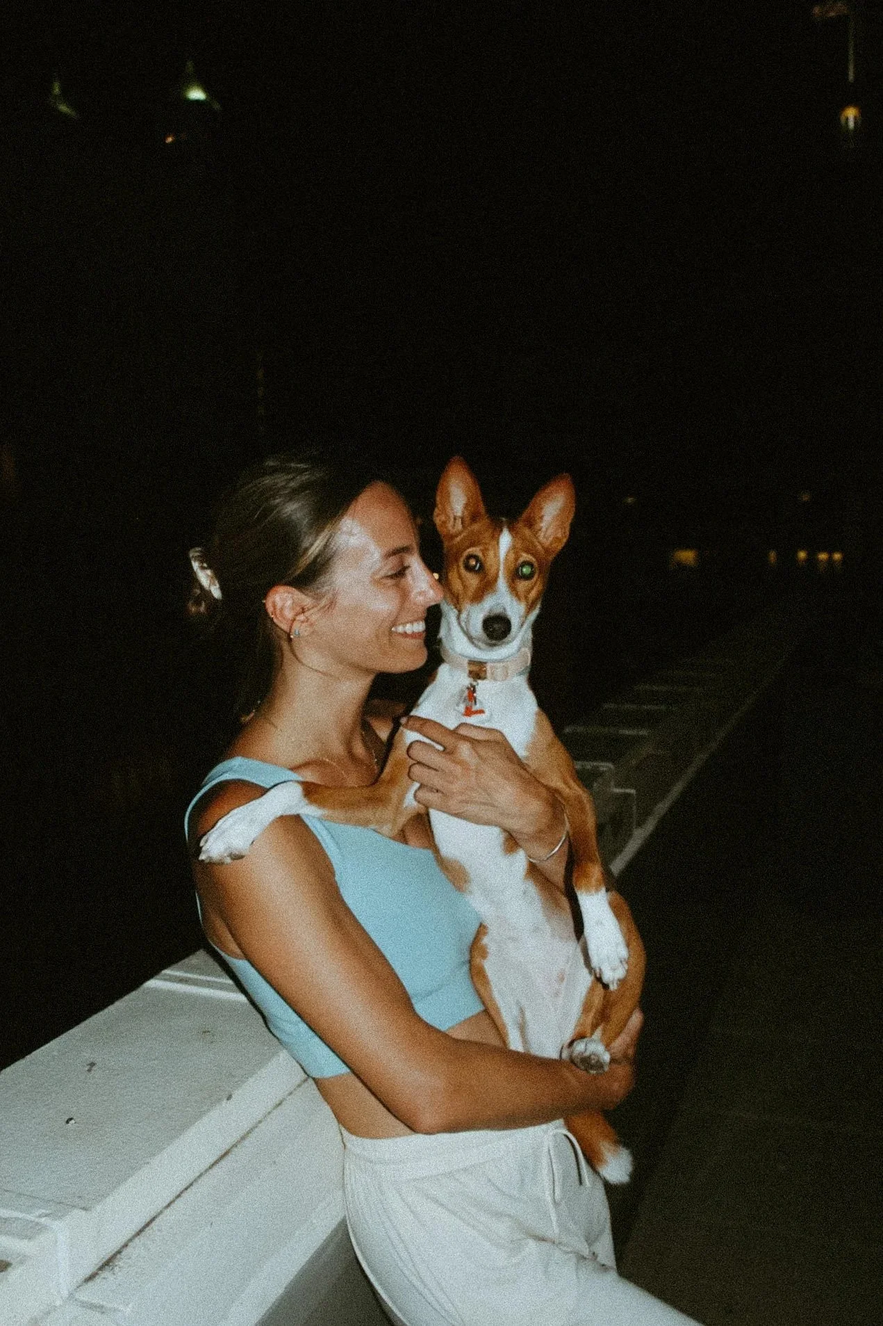 A woman in a light blue sleeveless top holding a small, brown and white dog with large ears and one green eye and one brown eye, at night outdoors.