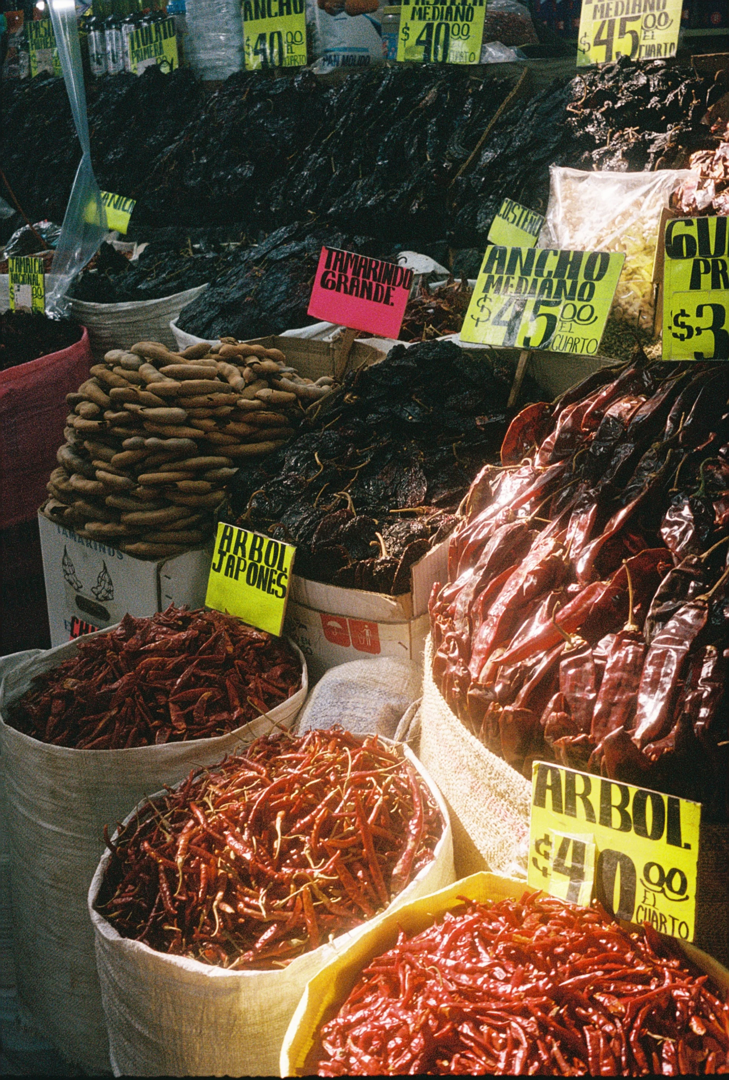 A market stall displaying various types of chili peppers in large sacks with bright yellow and pink signs indicating prices and types.