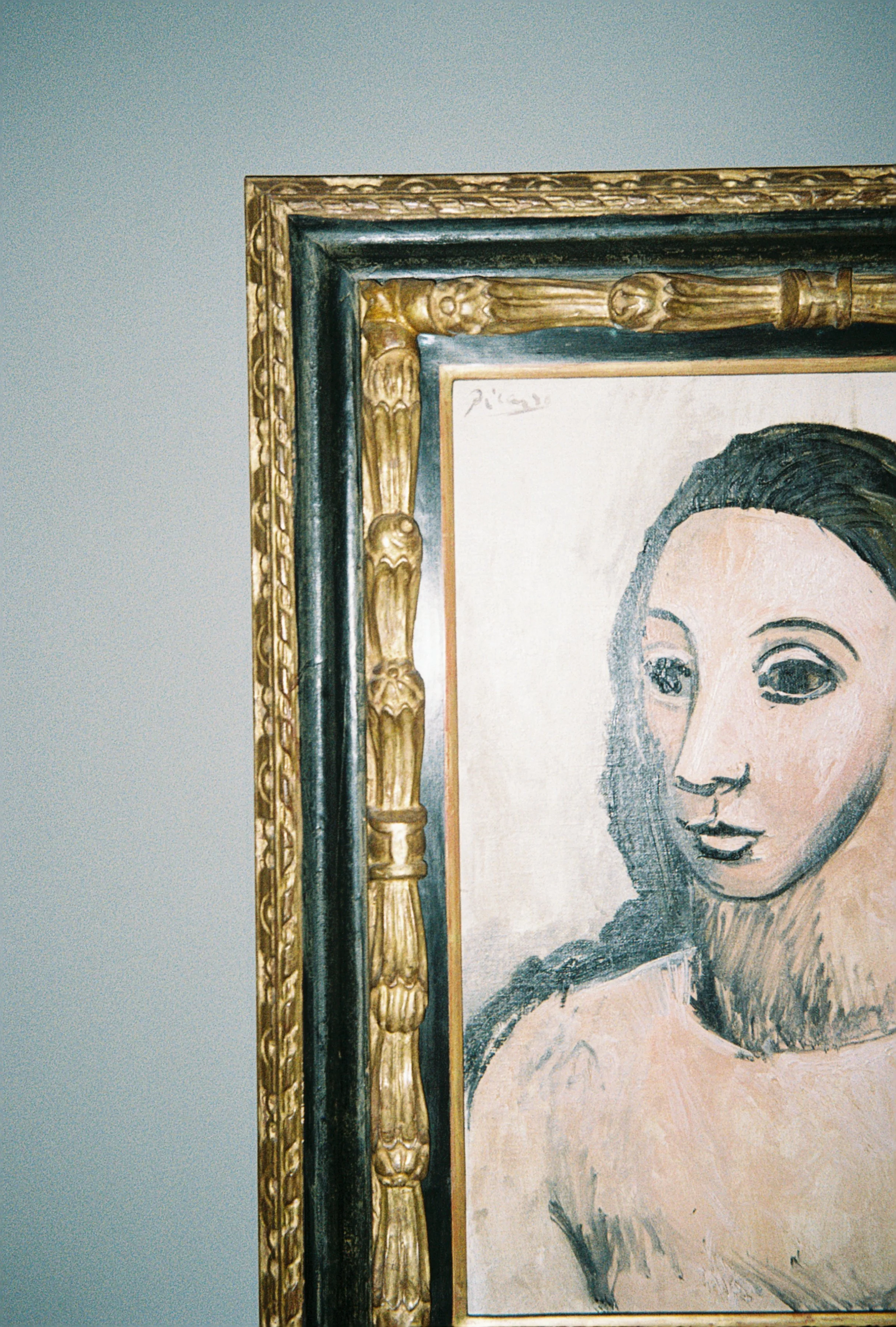 Close-up of the upper left corner of a framed painting of a woman's face, with visible gold ornate frame and signature area.