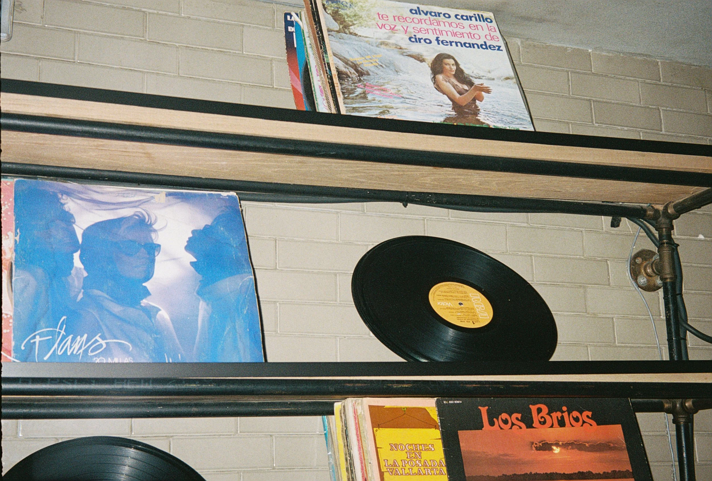 Shelf with vinyl records and magazines against a beige brick wall. Prominent magazine cover features a woman in water with text in Spanish.
