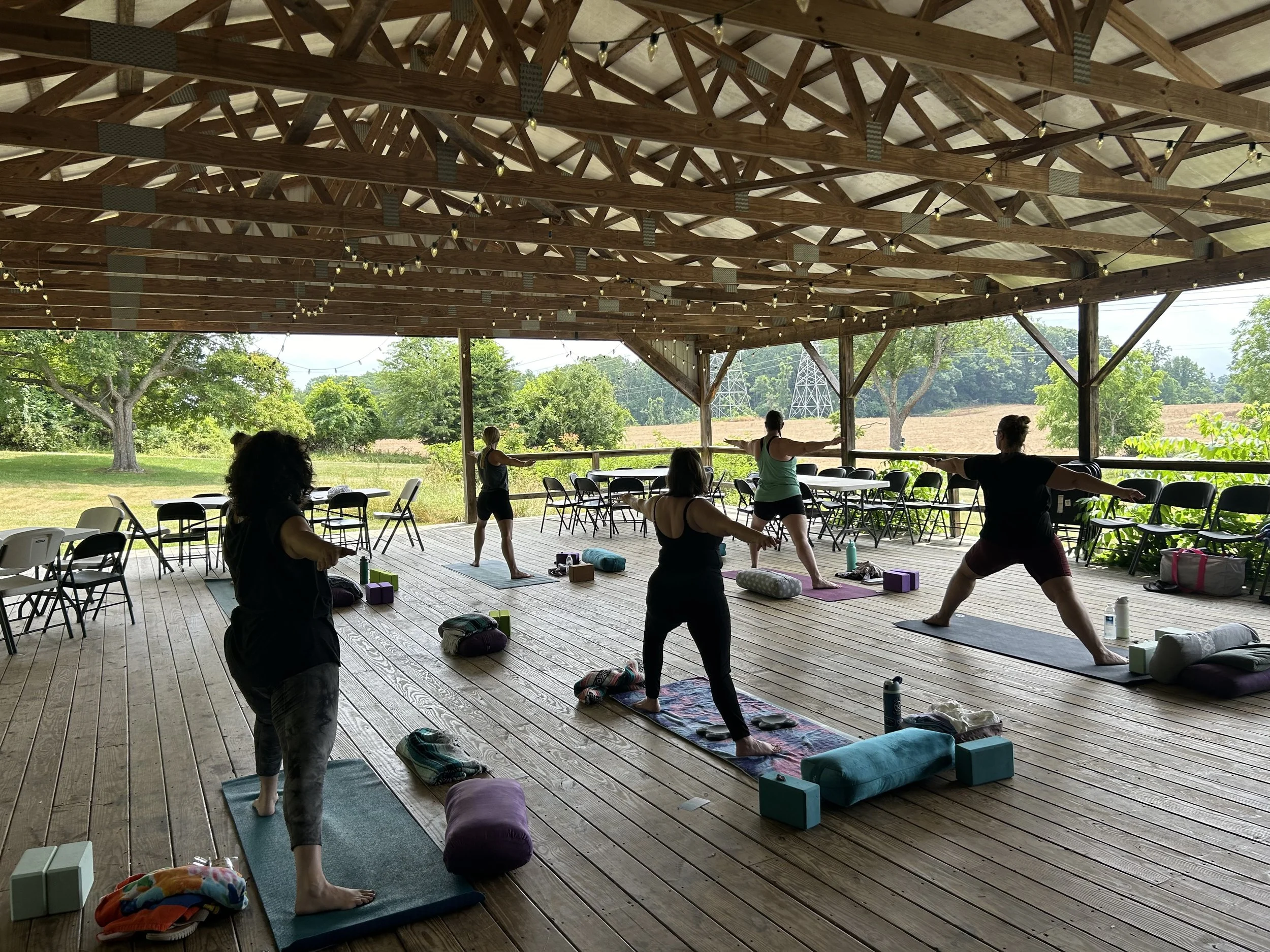 A group of people participating in a yoga class on an outdoor wooden pavilion with open sides, surrounded by trees and greenery.