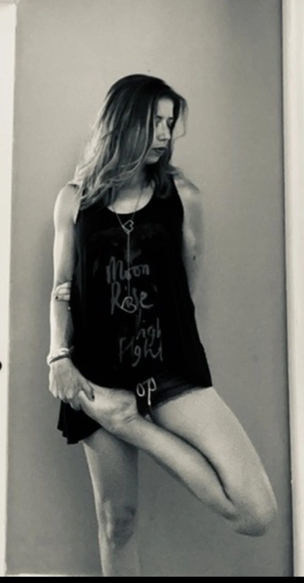 A woman with wavy hair standing against a plain wall, wearing a tank top, shorts, and fishnet stockings, looking down.