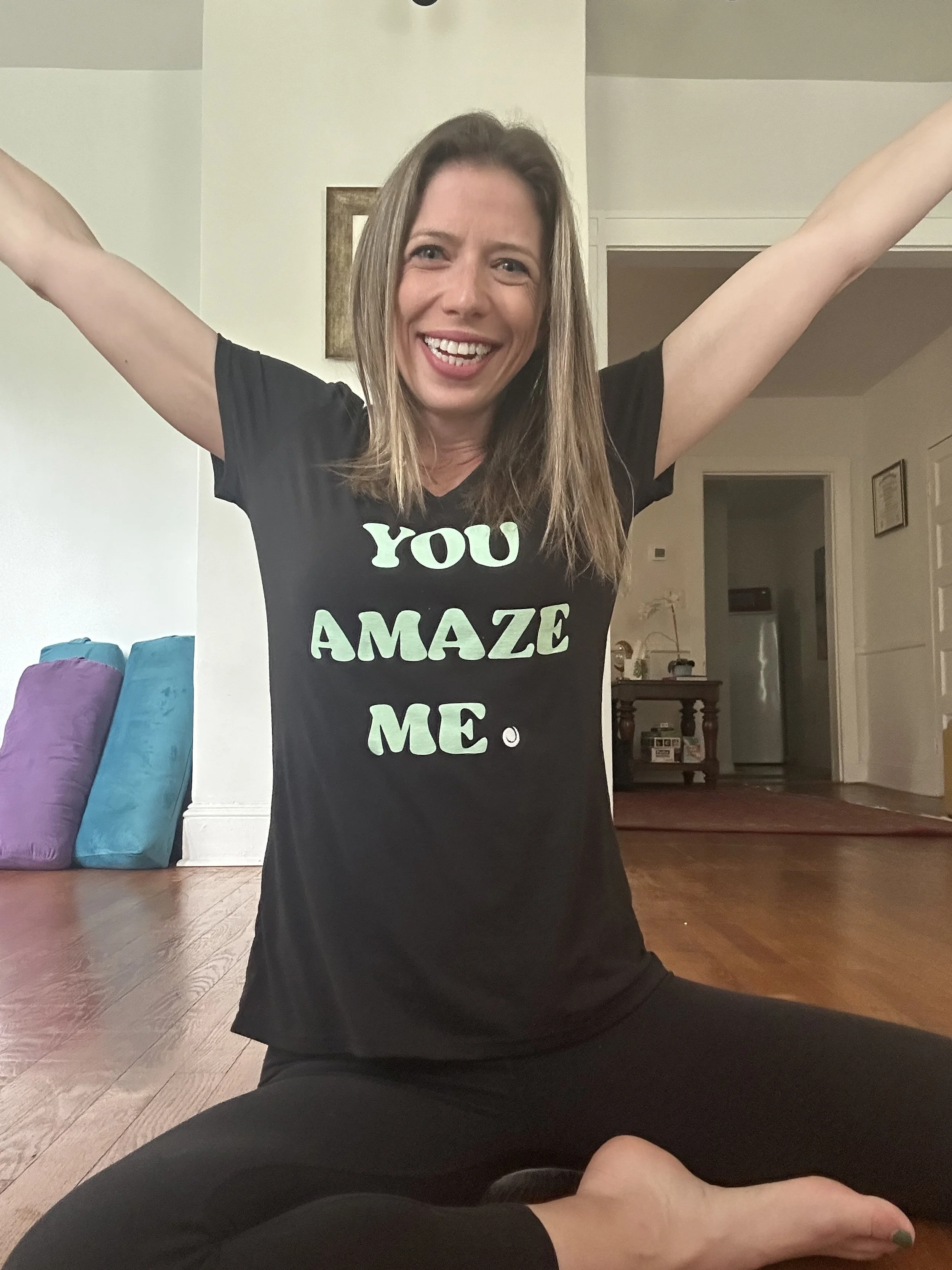 A woman sitting on the floor in a yoga pose with arms raised, smiling, wearing a black T-shirt that says 'YOU AMAZE ME.'