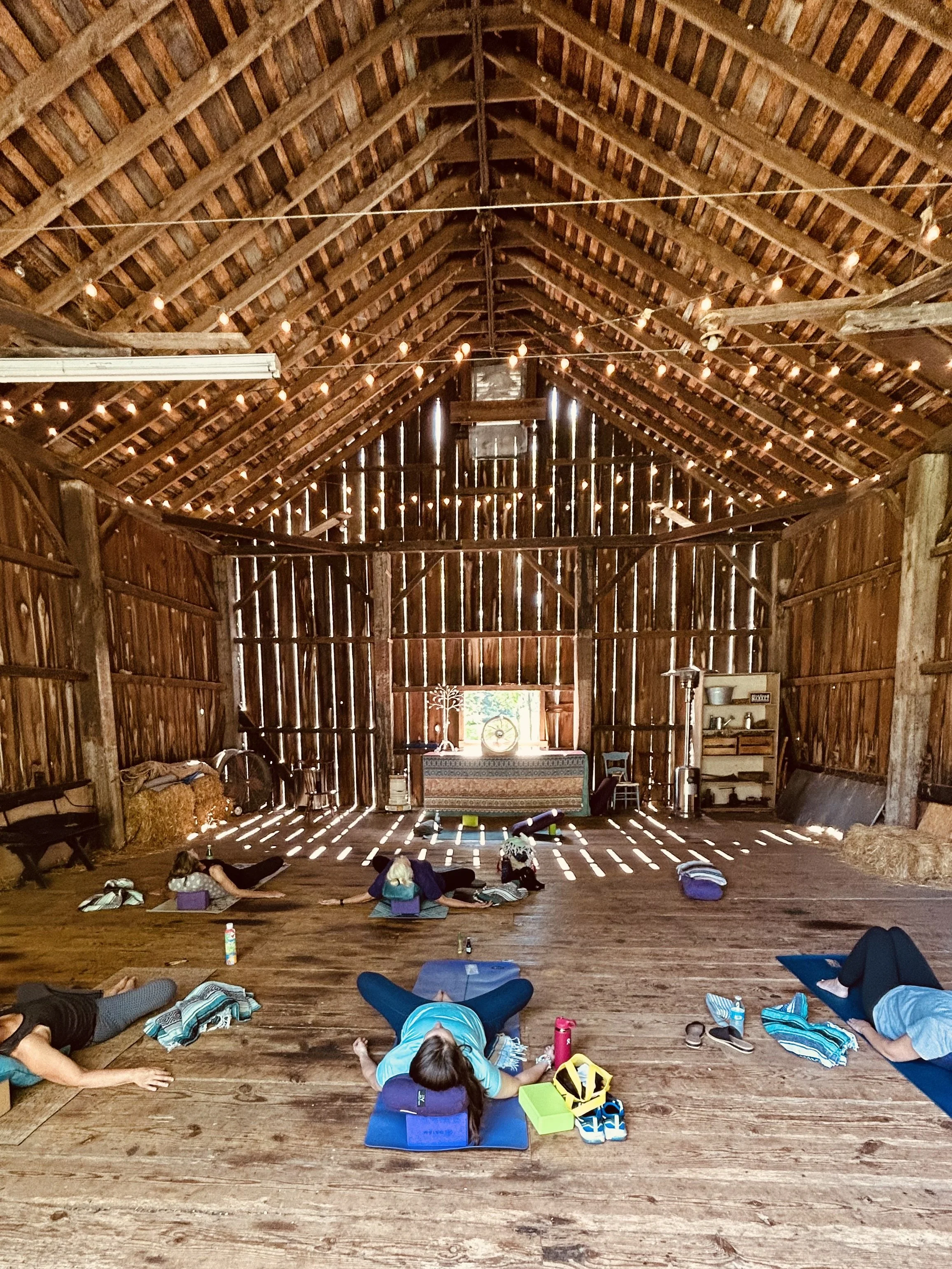People participating in a yoga class inside a rustic barn with string lights, practicing poses on mats with personal belongings around.