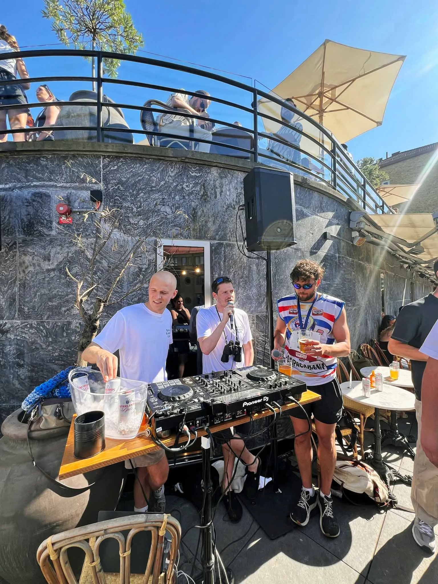A DJ setup outdoors with three men, two holding drinks, and one speaking into a microphone, under bright sunlight with parasols and tables around.