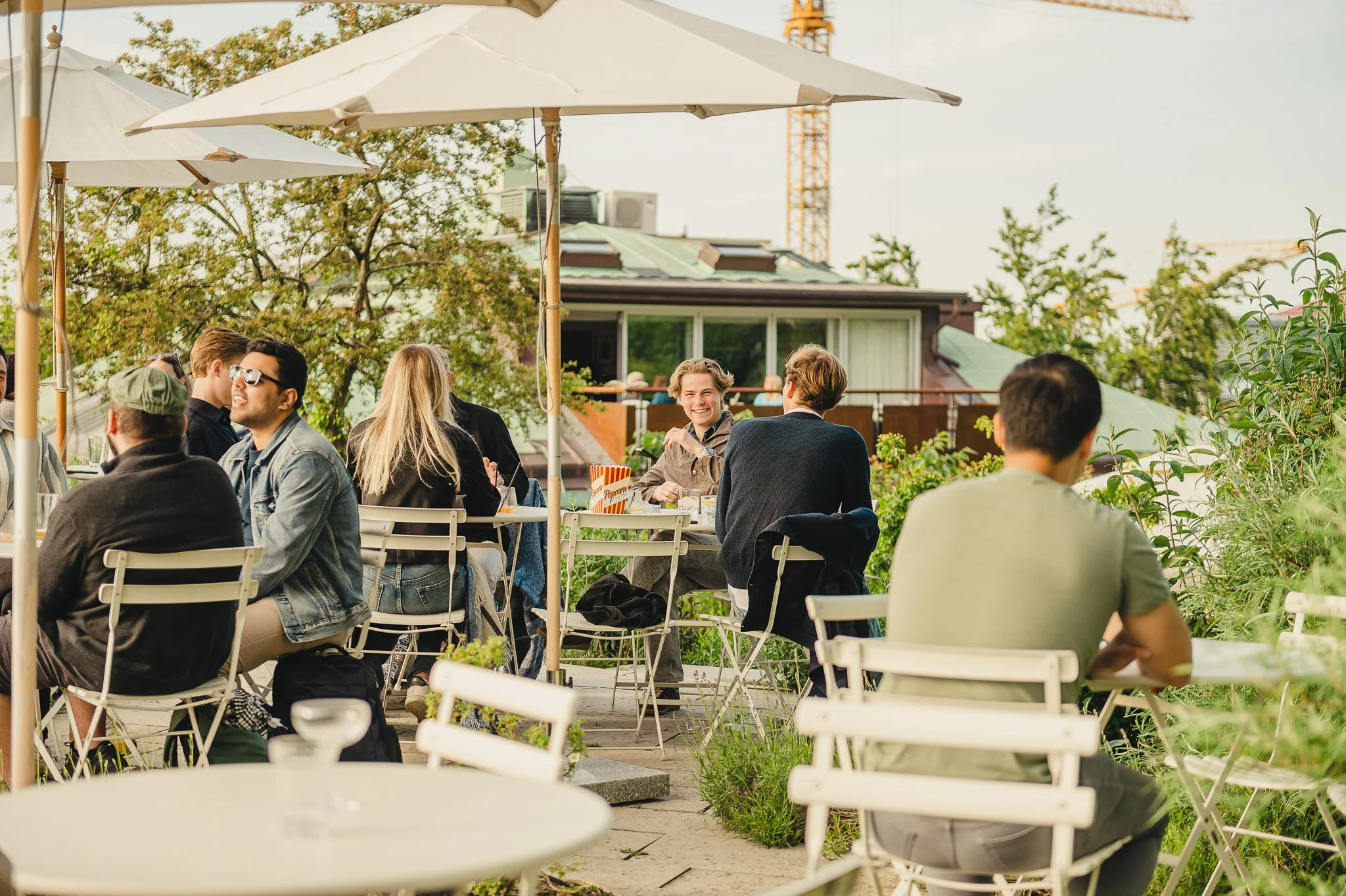 People sitting at outdoor tables on a patio enjoying conversations and drinks, shaded by large umbrellas, with greenery and buildings in the background.
