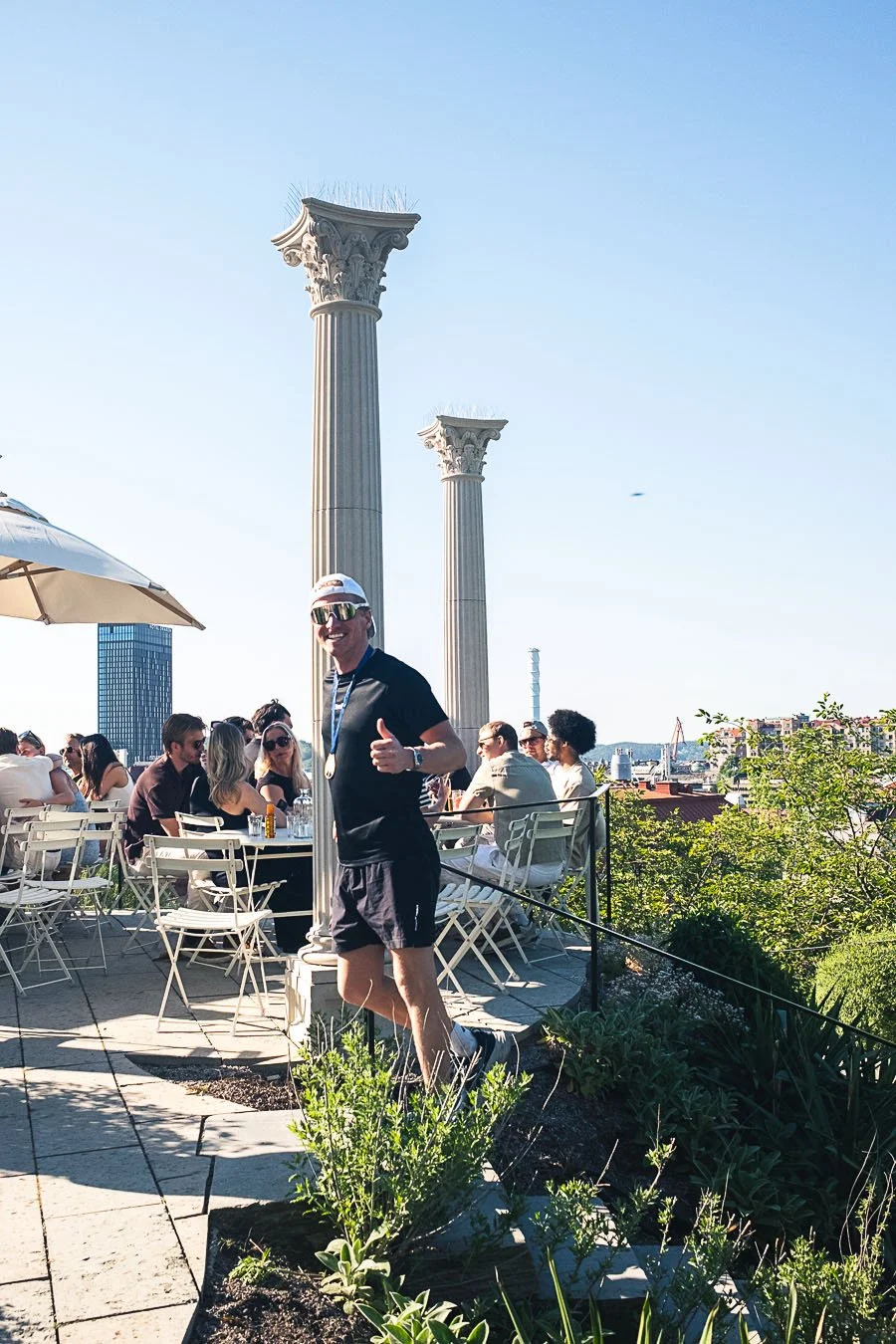 A man in black athletic clothing and sunglasses giving a thumbs-up during a daytime outdoor gathering with ancient Greek-style columns in the background.