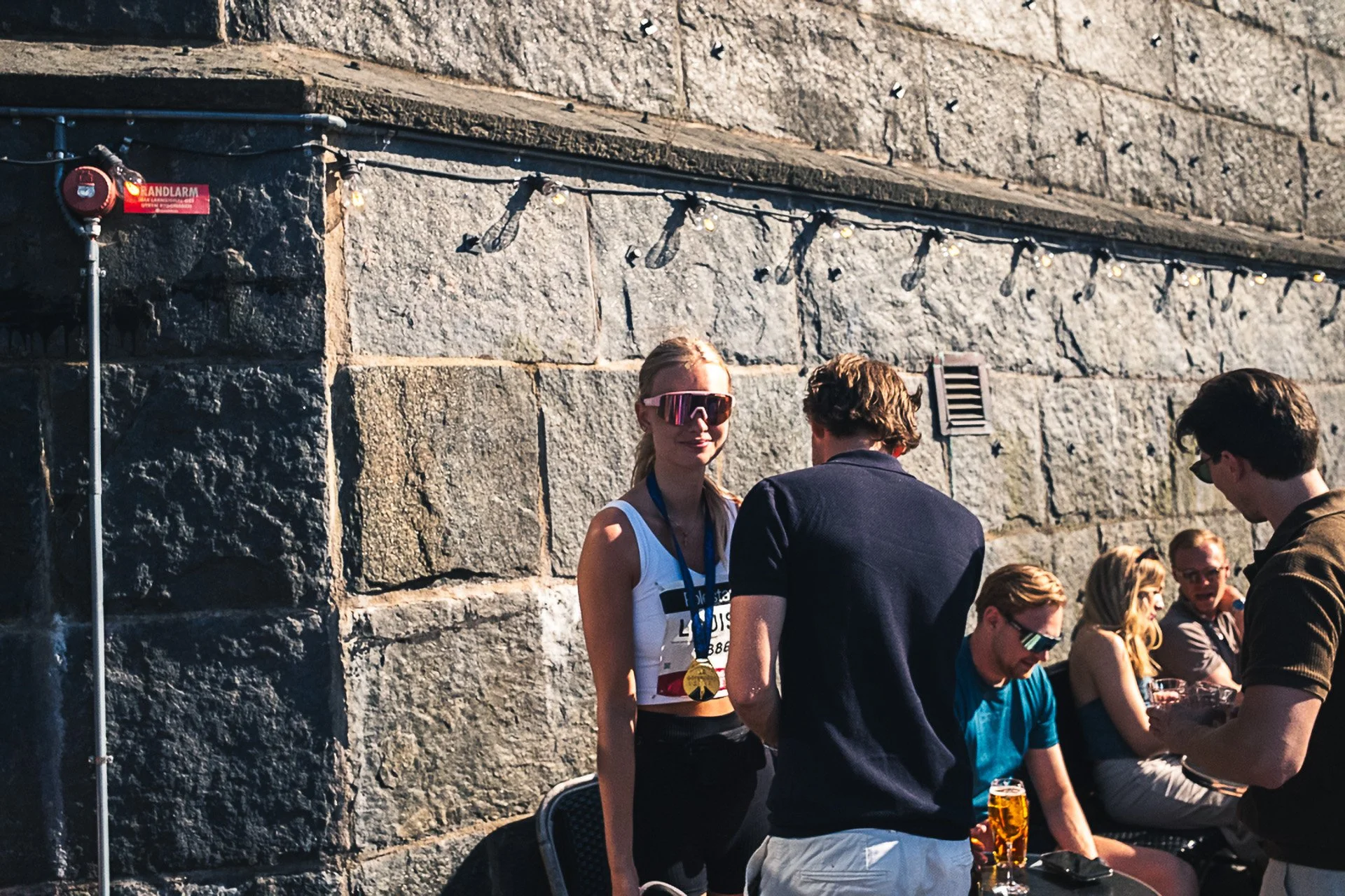 People gathered outdoors near a stone wall, some wearing sunglasses and athletic clothing; one person has a medal around their neck.