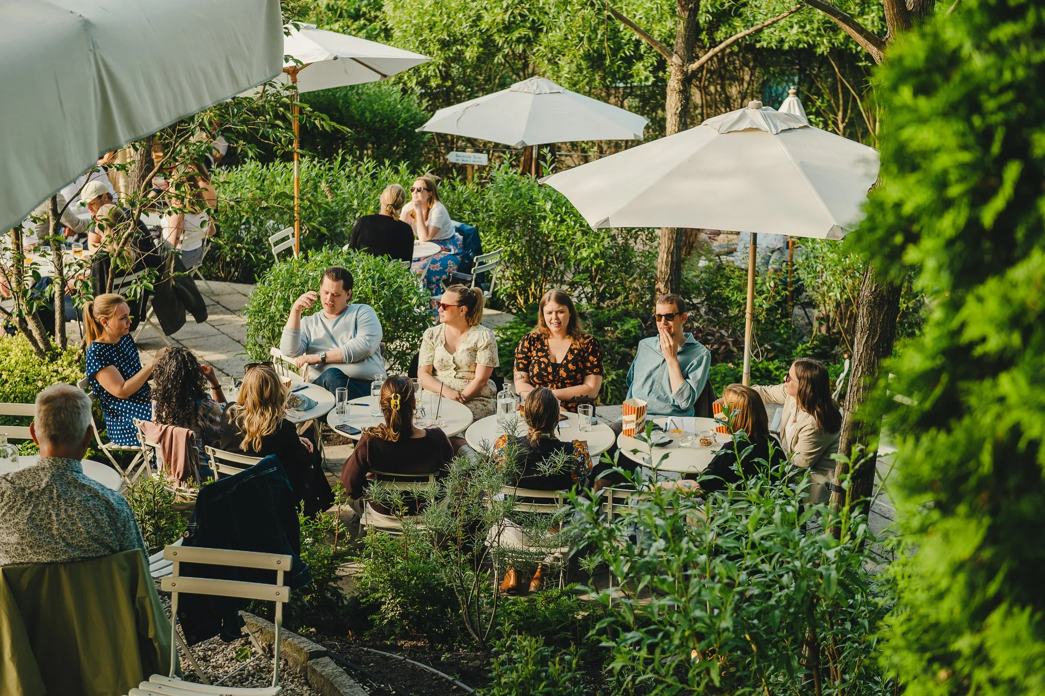 People sitting at outdoor tables under large white umbrellas at a green garden restaurant on a sunny day.