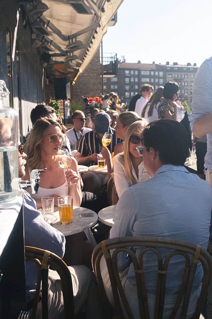 People sitting at outdoor tables on a rooftop patio, enjoying drinks in sunlight with city buildings in the background.