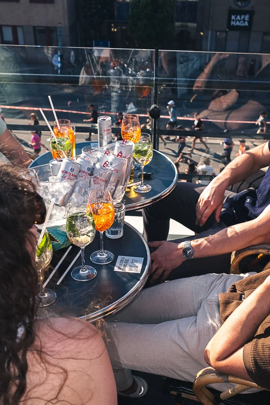 People sitting at an outdoor café table with drinks, overlooking a busy city street with pedestrians.