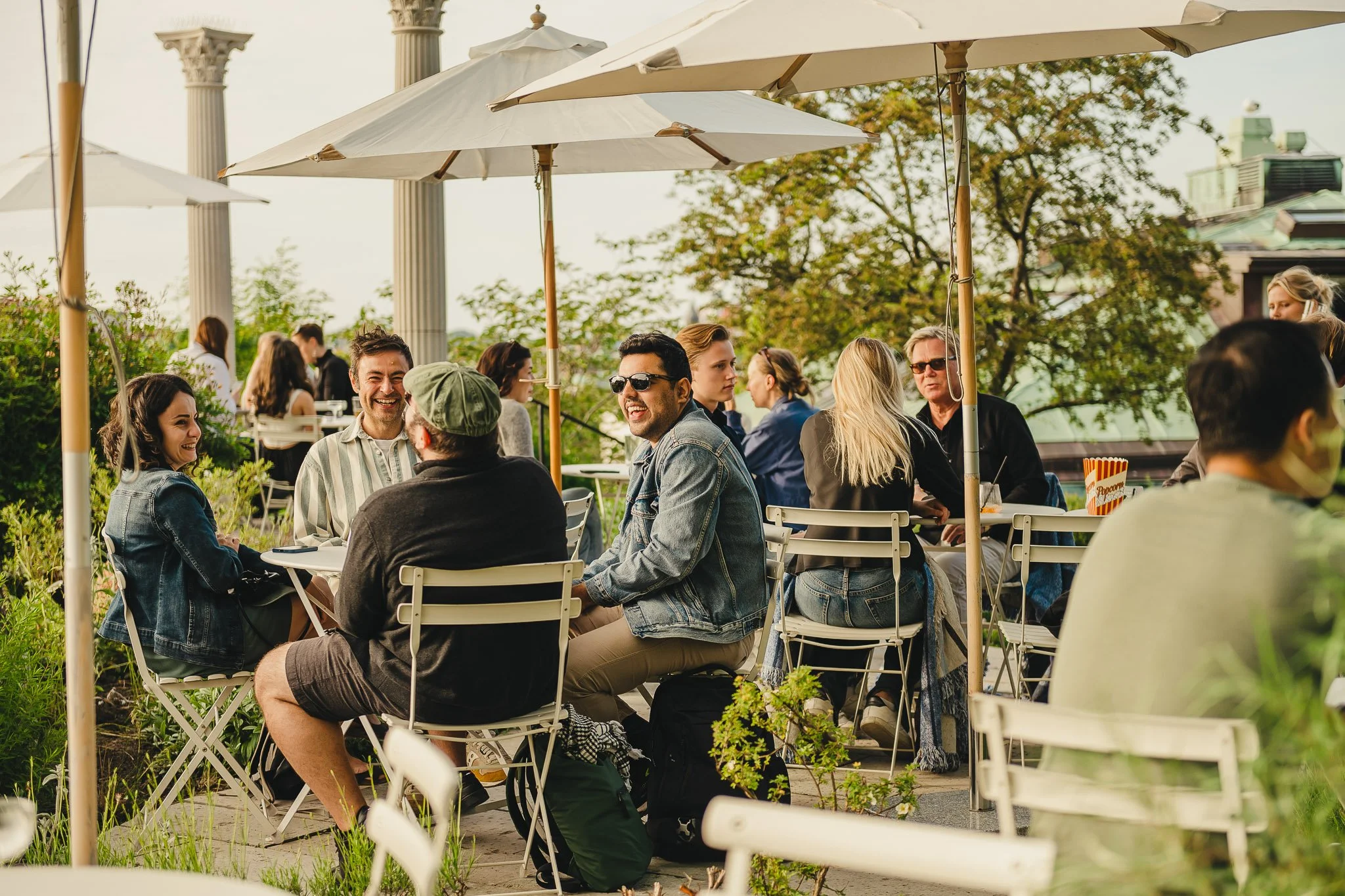 People sitting at tables outdoors enjoying a sunny day at a cafe, some wearing sunglasses, under large umbrellas with trees and columns in the background.